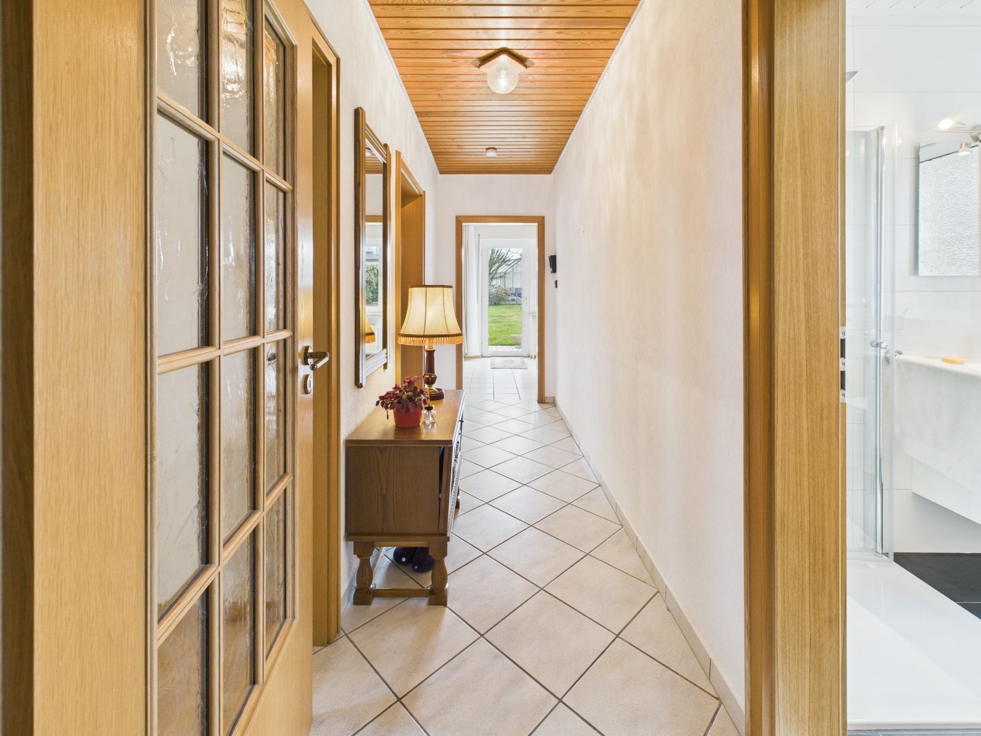 Heller Flur mit Fliesenboden, Holzdecke, Kommode mit Lampe und Spiegel an der Wand.