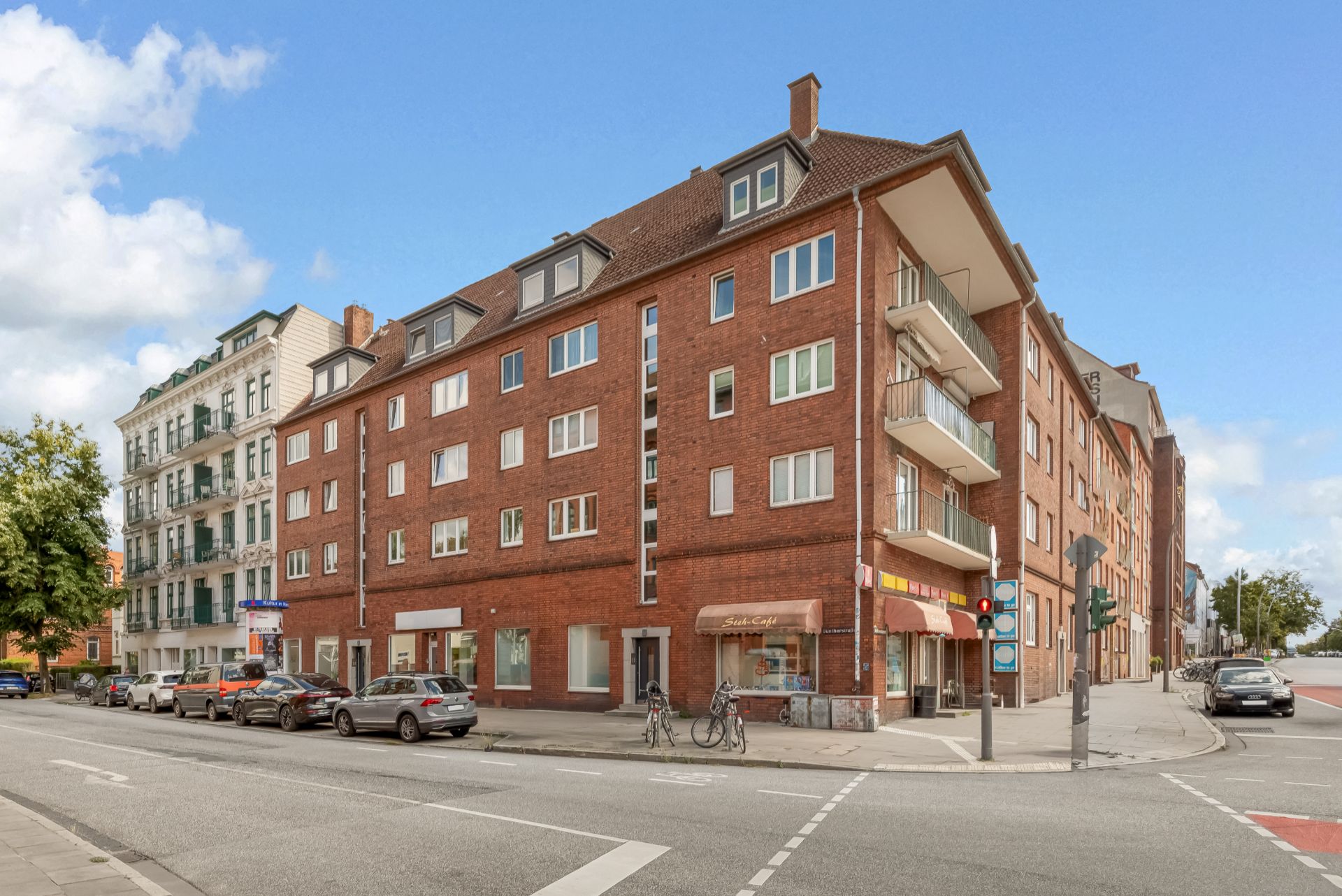 Mehrstöckiges rotes Wohngebäude mit Balkonen an einer Straßenecke, mehrere geparkte Autos und Fahrräder.