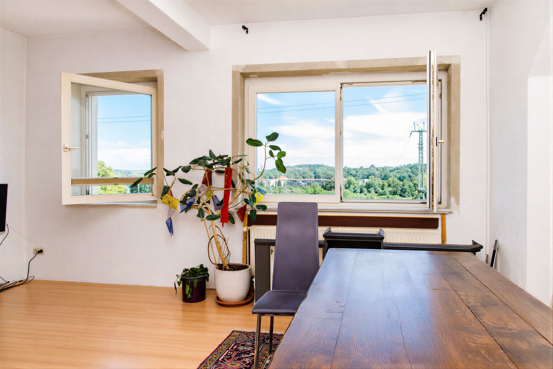 Helles Zimmer mit Holzfußboden, offenem Fenster, Schreibtisch, Stuhl und Topfpflanzen.