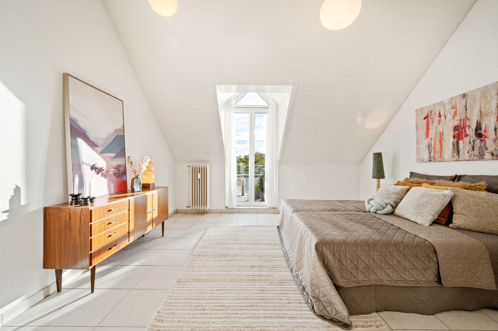 Helles Schlafzimmer mit Doppelbett, Kommode, großem Fenster und dekorativen Bildern an den Wänden.