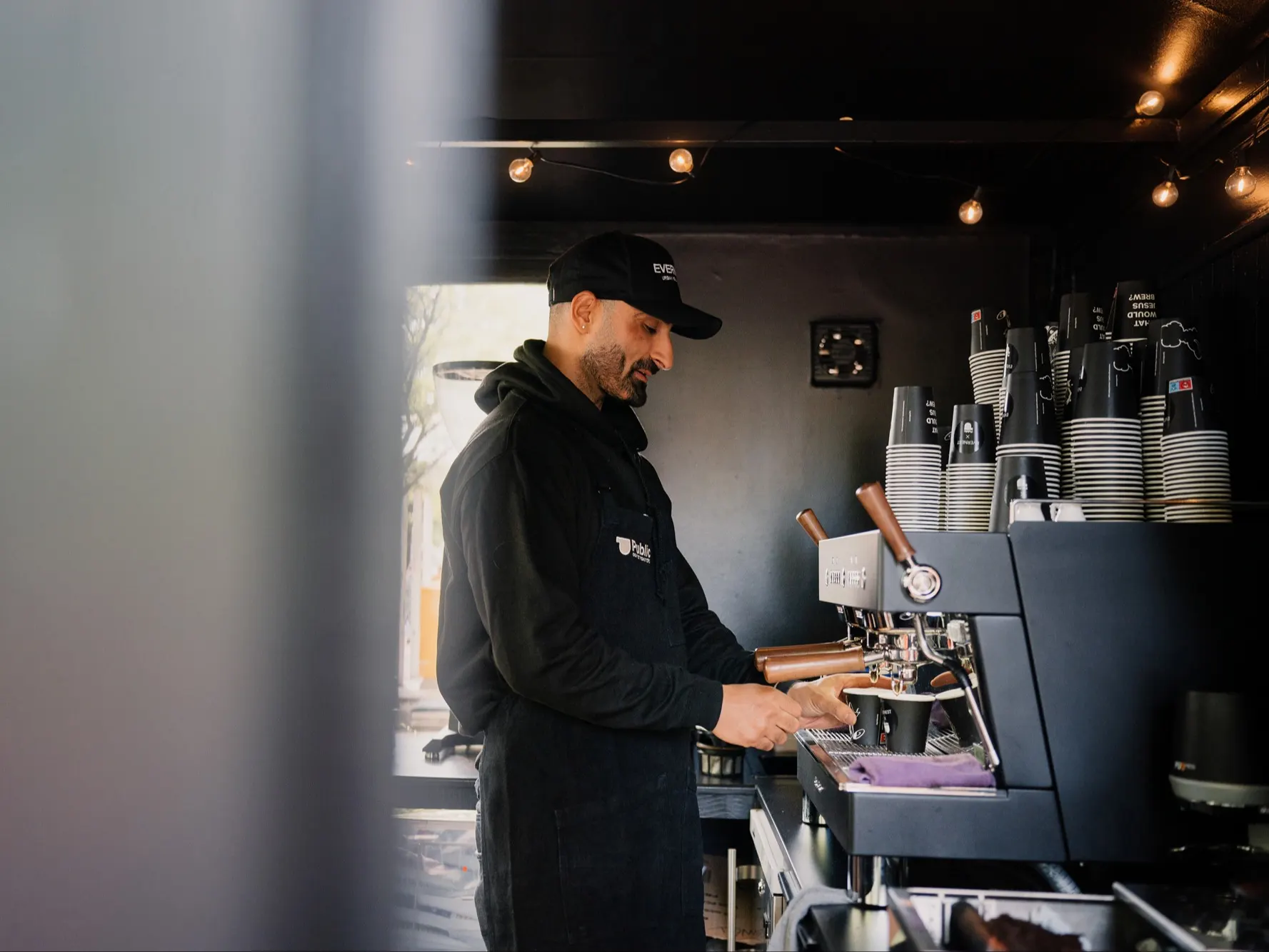 Mann bereitet an einer Kaffeemaschine einen Kaffee zu, viele Becher sind gestapelt.