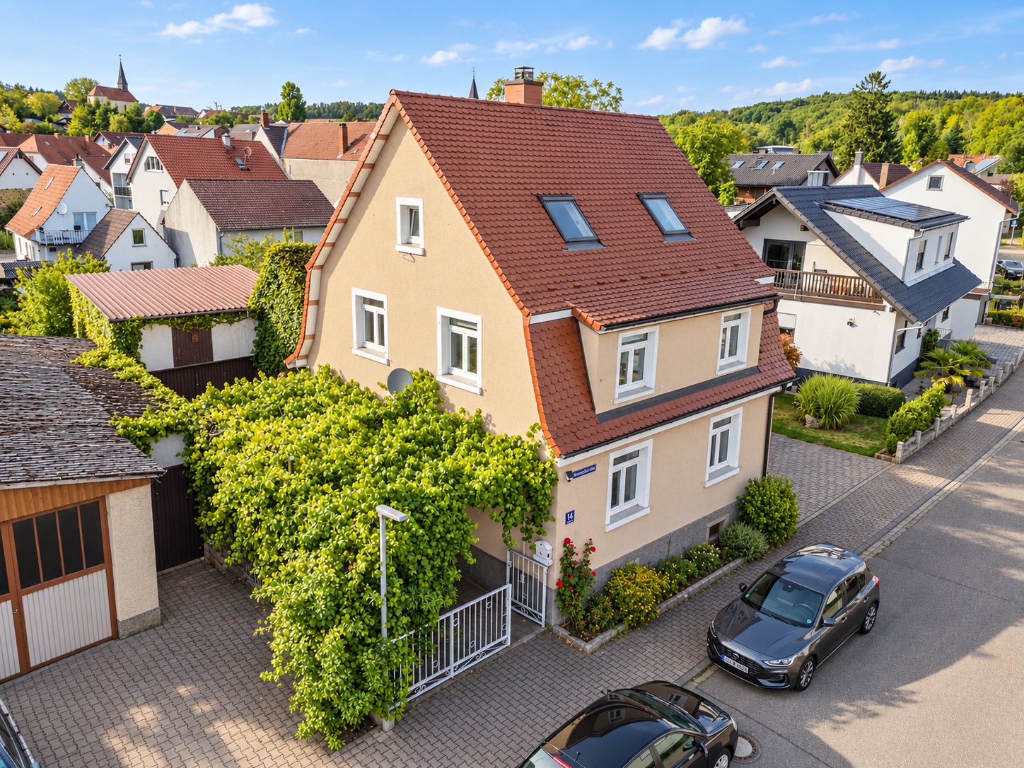 Zweistöckiges Wohnhaus mit rotem Dach, bewachsener Eingang und zwei geparkte Autos an der Straße.