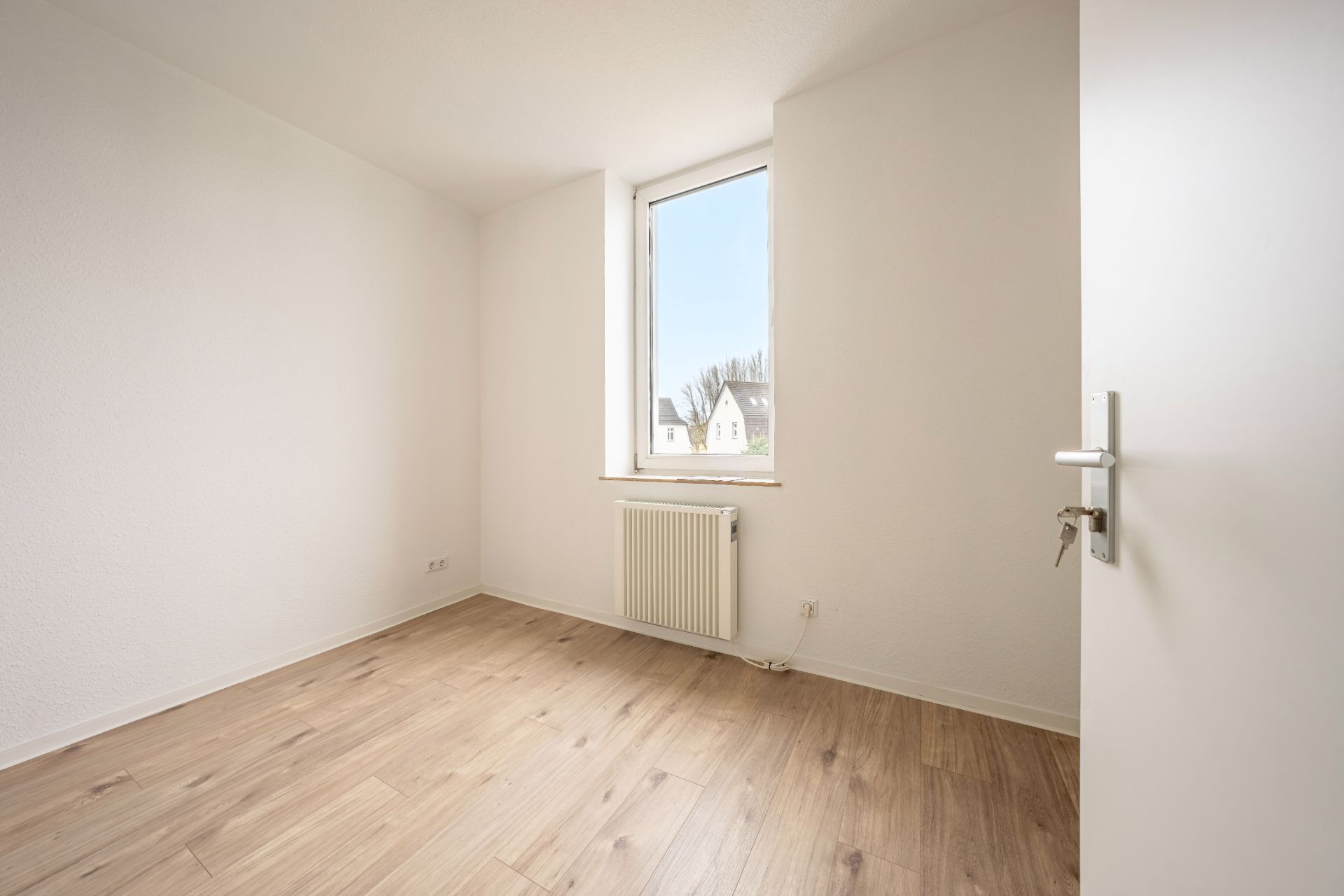 Helles, leeres Zimmer mit Holzfußboden, weißer Wand, Fenster und Heizkörper.
