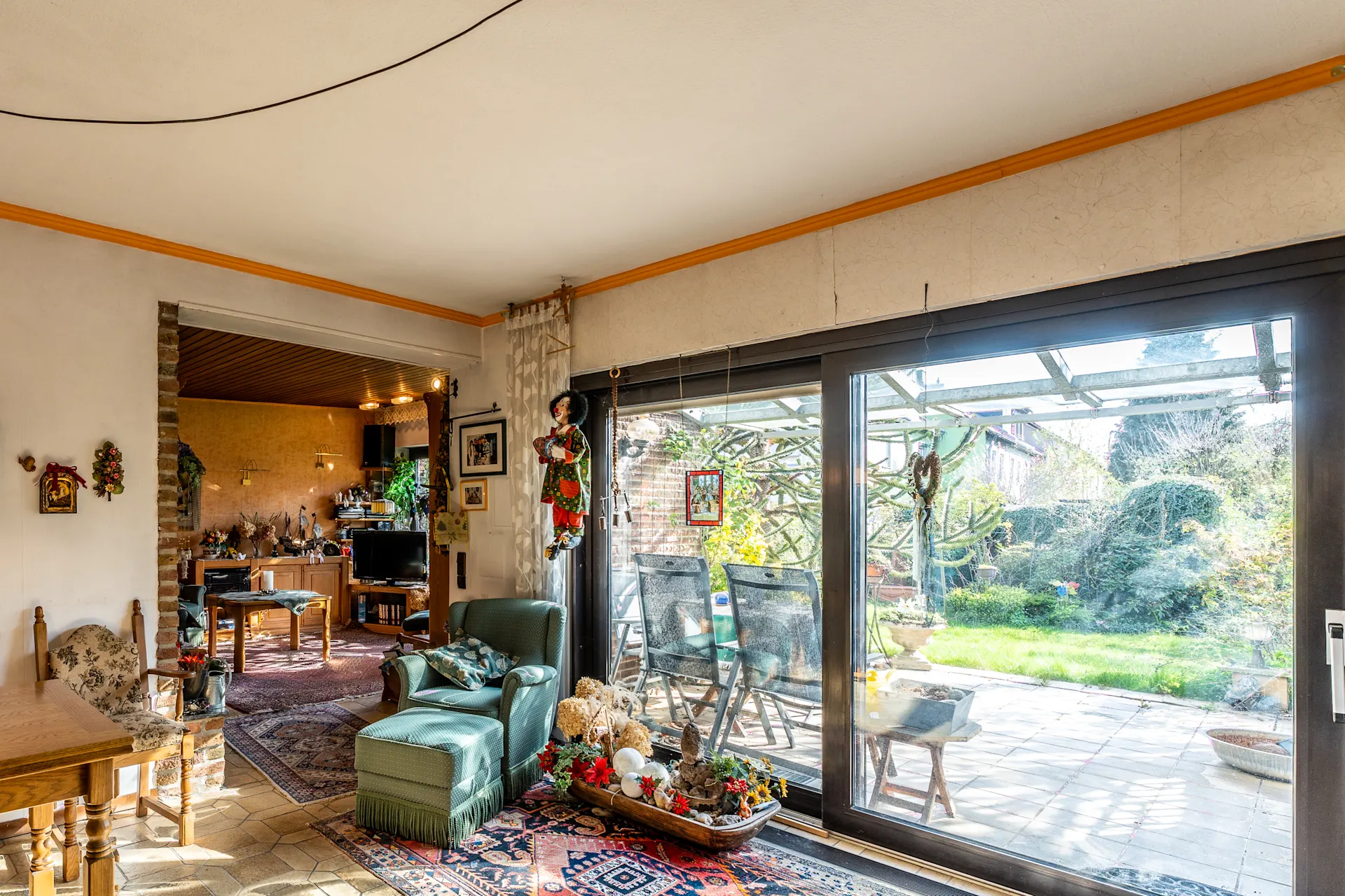 Helles Wohnzimmer mit großen Fenstern, Blick auf Terrasse und Garten, Sessel und Dekorationen sichtbar.