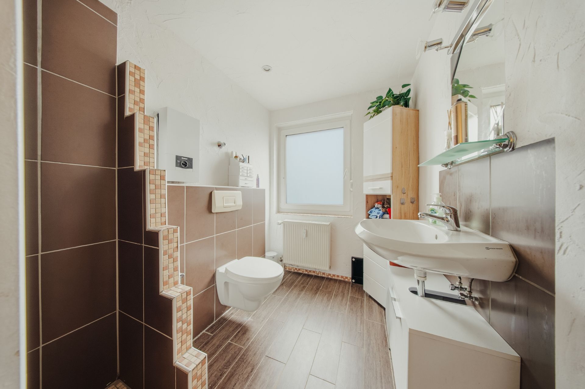 Modernes Badezimmer mit braunen Fliesen, Wand-WC, Waschbecken und Fenster.