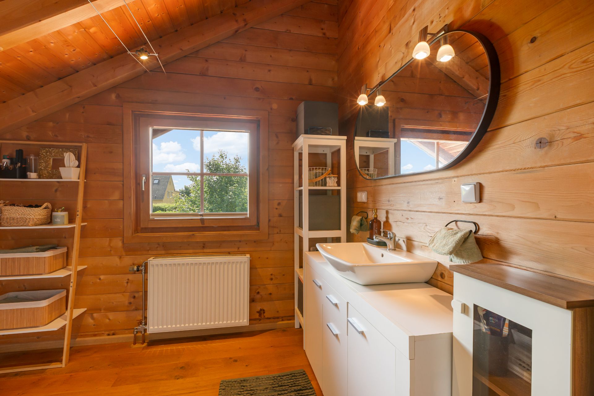 Holz-Badezimmer mit Waschbecken, rundem Spiegel, Regal und Fenster mit Ausblick.