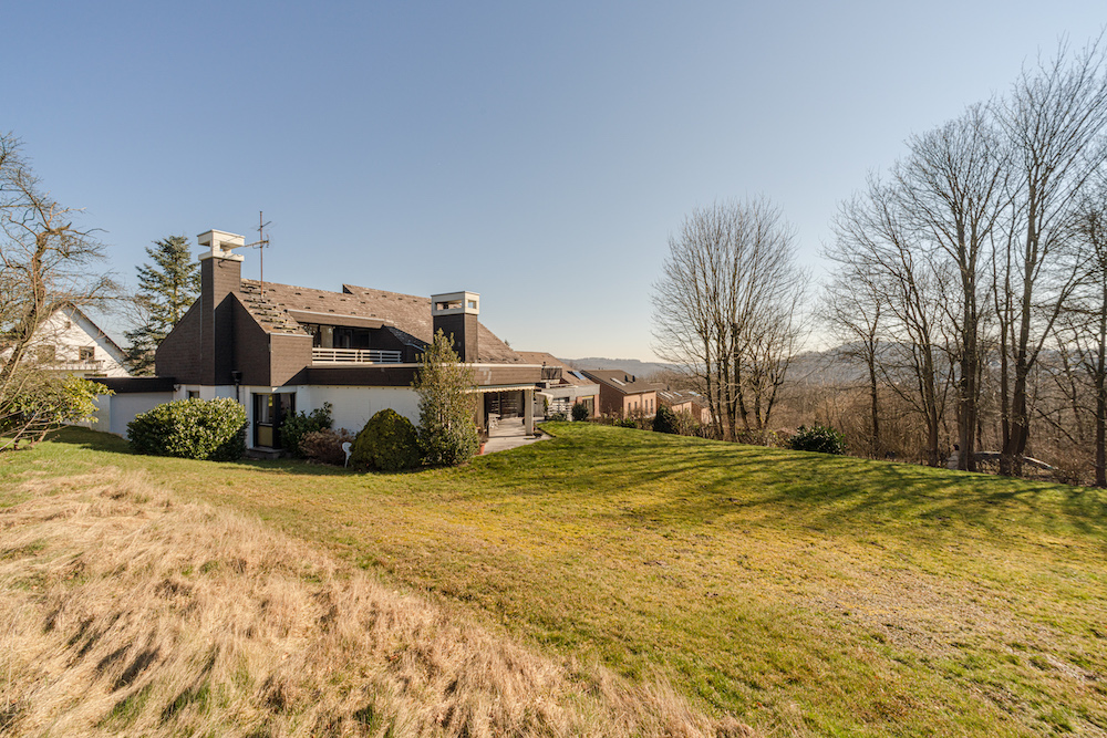 Modernes Wohnhaus mit großem Garten und Bäumen, im Hintergrund weite Landschaft.