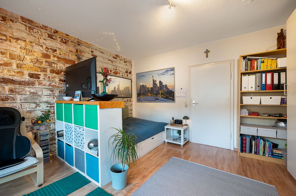 Wohnzimmer mit Bett, Regal, Schreibtisch, Sessel und gemauerter Wand.