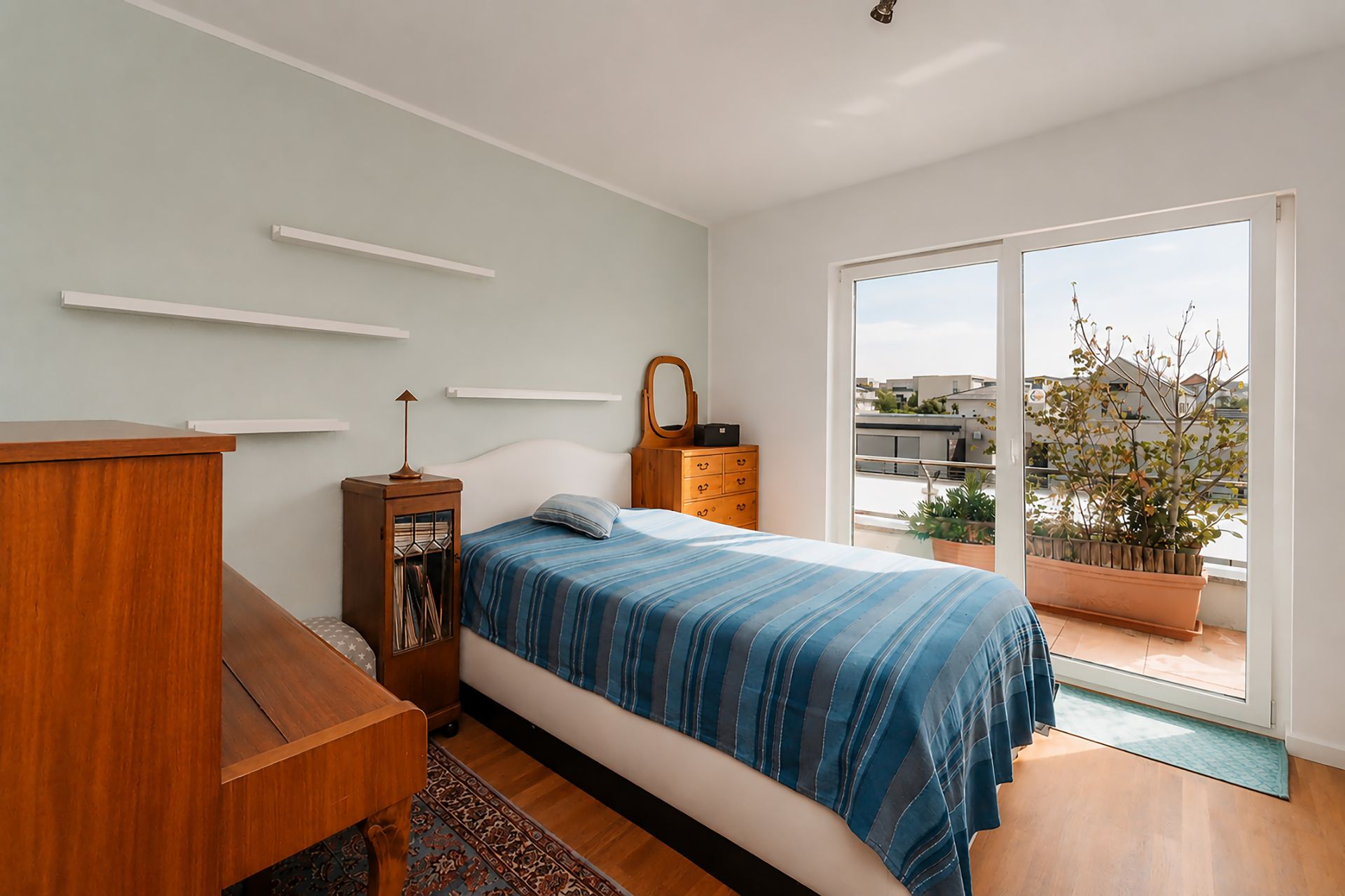Helles Schlafzimmer mit Einzelbett, Holzmöbeln und Zugang zu einem Balkon mit Pflanzen.