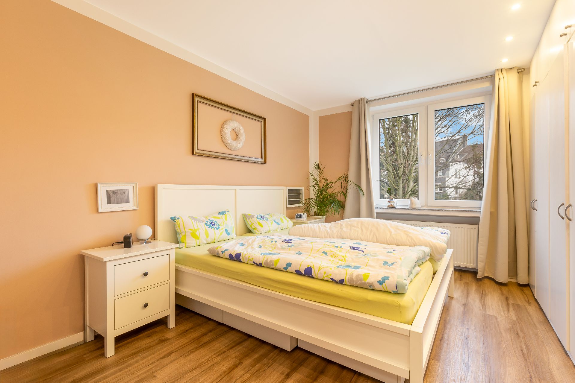 Helles Schlafzimmer mit Doppelbett, Nachttisch, Pflanzen und großem Fenster.