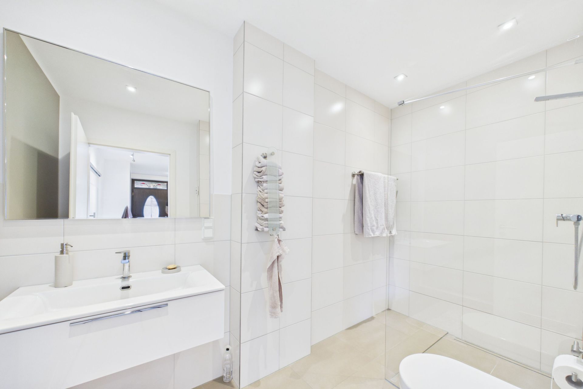 Modernes, weiß gefliestes Badezimmer mit Dusche, Waschbecken, Spiegel und Handtüchern.