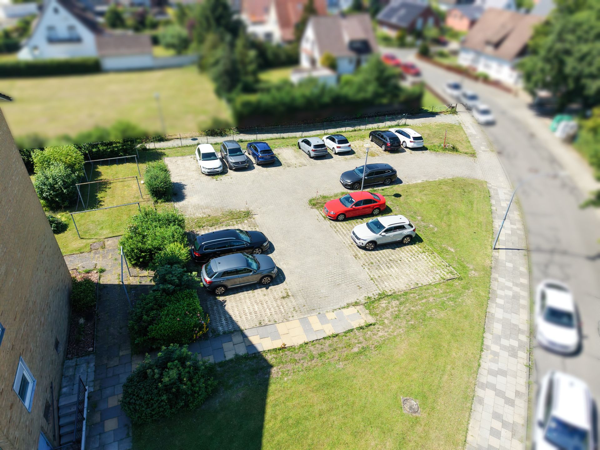 Parkplatz mit mehreren Autos neben einem Wohnhaus, umgeben von Wiese und Gehweg.