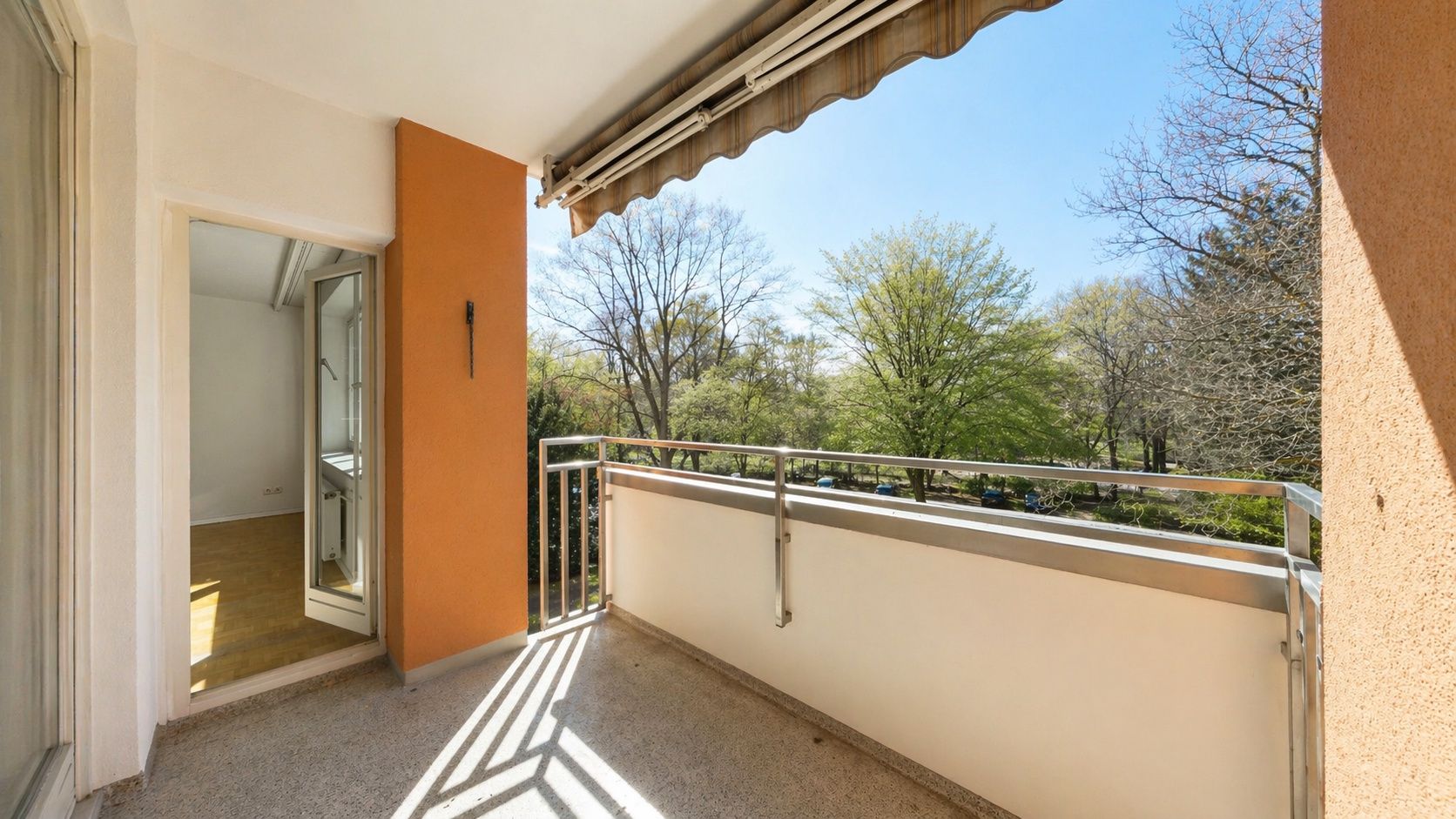 Ein sonniger Balkon mit Geländer, Blick auf Bäume, Markise und Zugang zu einem Zimmer.