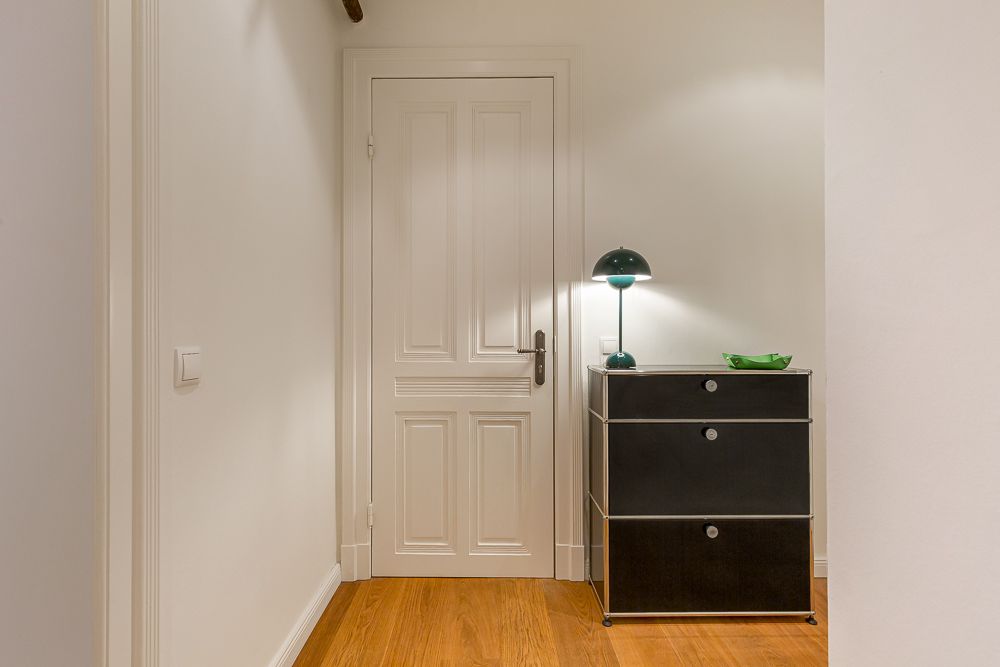 Flur mit weißer Tür, schwarzer Kommode, Tischlampe und Holzfußboden.