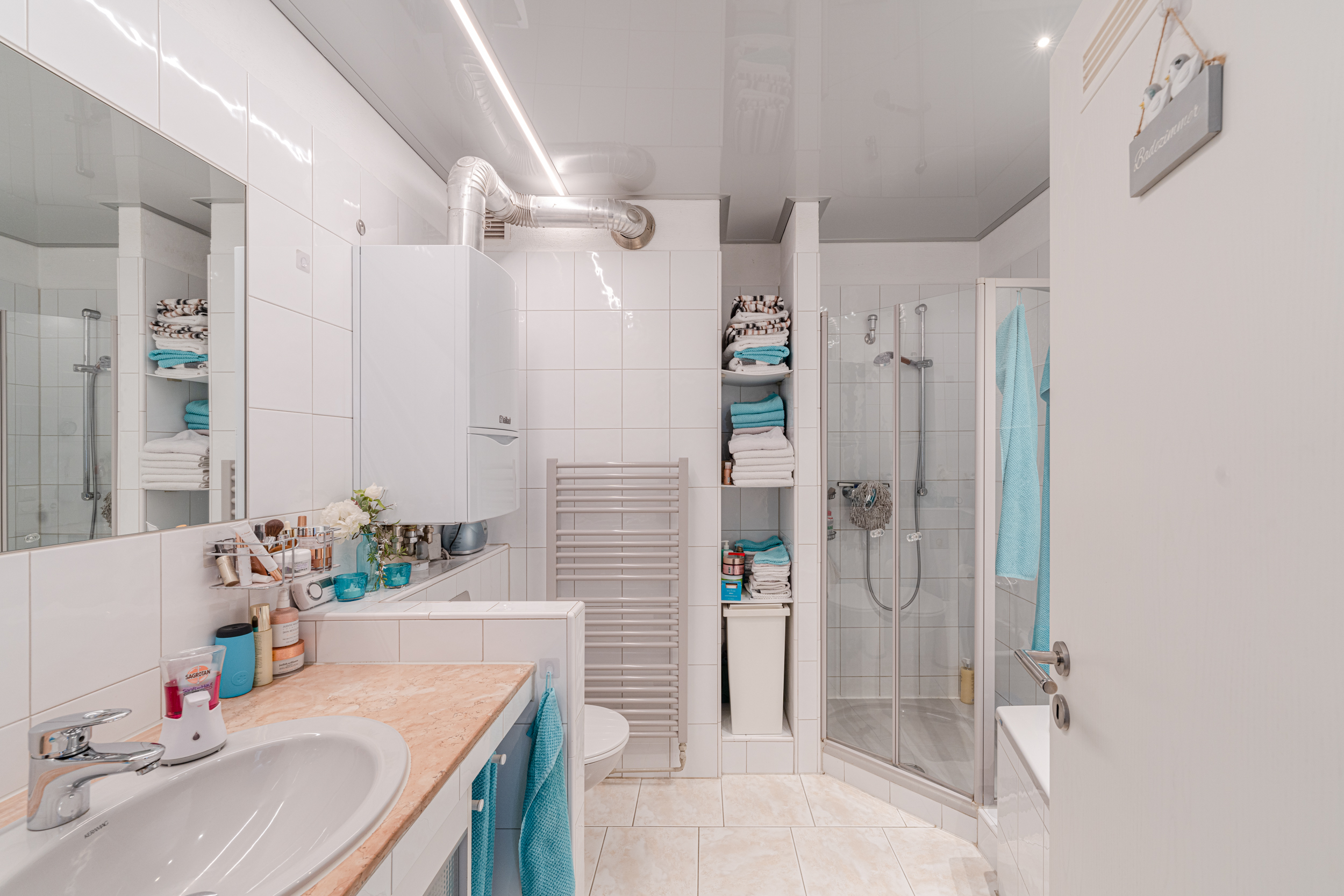 Helles Badezimmer mit Dusche, Waschbecken, Handtuchtrockner und Regal mit Handtüchern.