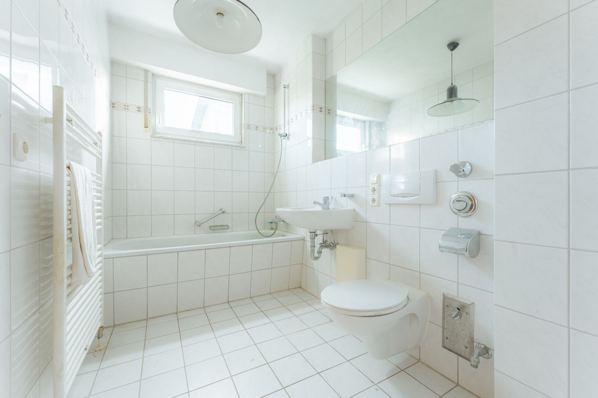 Helles Badezimmer mit Badewanne, WC, Waschbecken, Spiegel und weiß gefliesten Wänden und Boden.