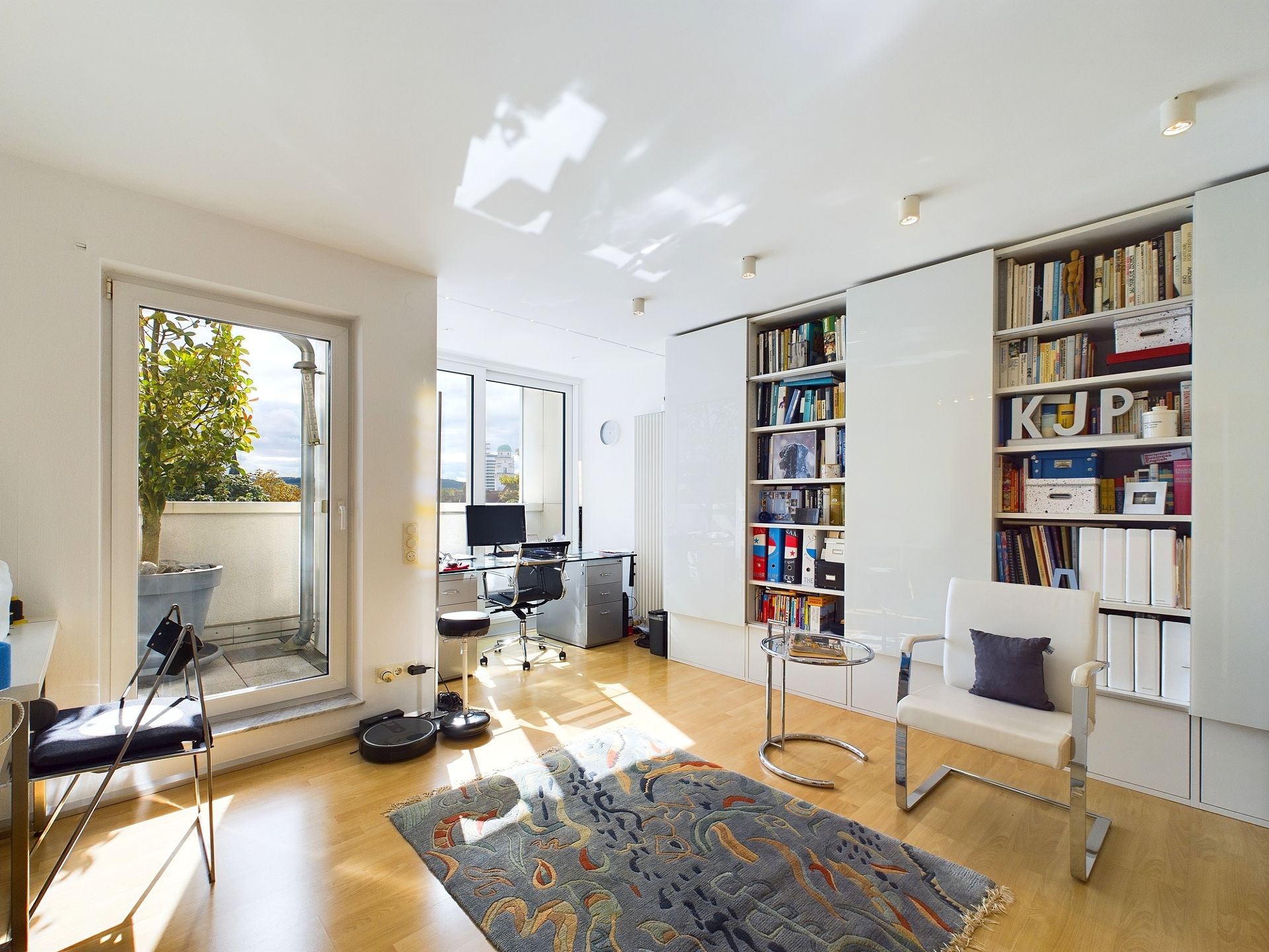 Helles Arbeitszimmer mit Regalen, Schreibtisch, Stuhl, Teppich und Zugang zu einem Balkon.