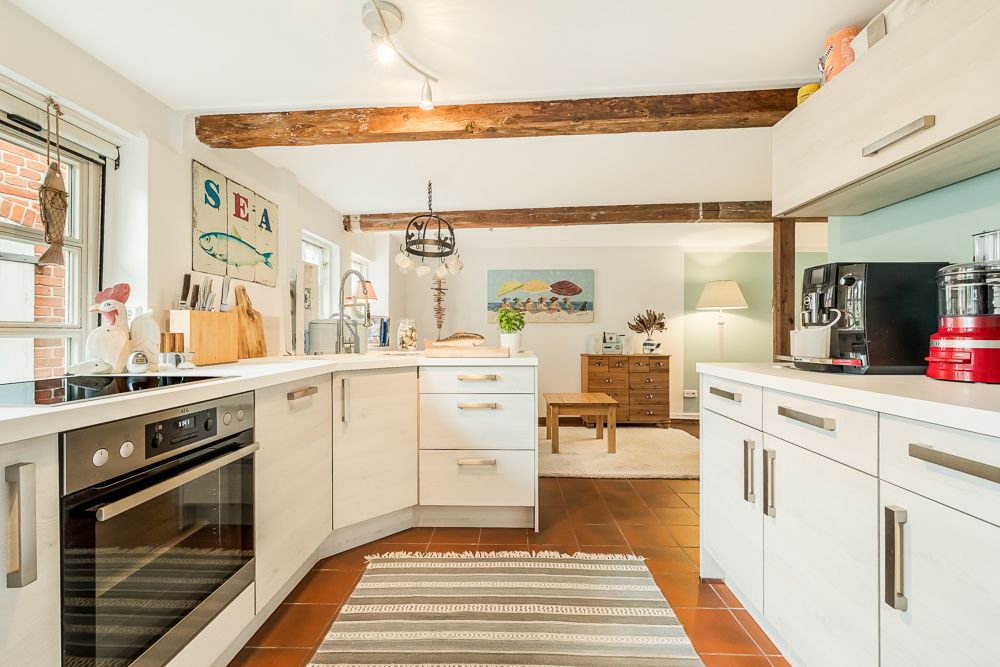 Helle Küche mit weißen Schränken und Holzbalken, angrenzend an ein Wohnzimmer mit Teppich und Tisch.