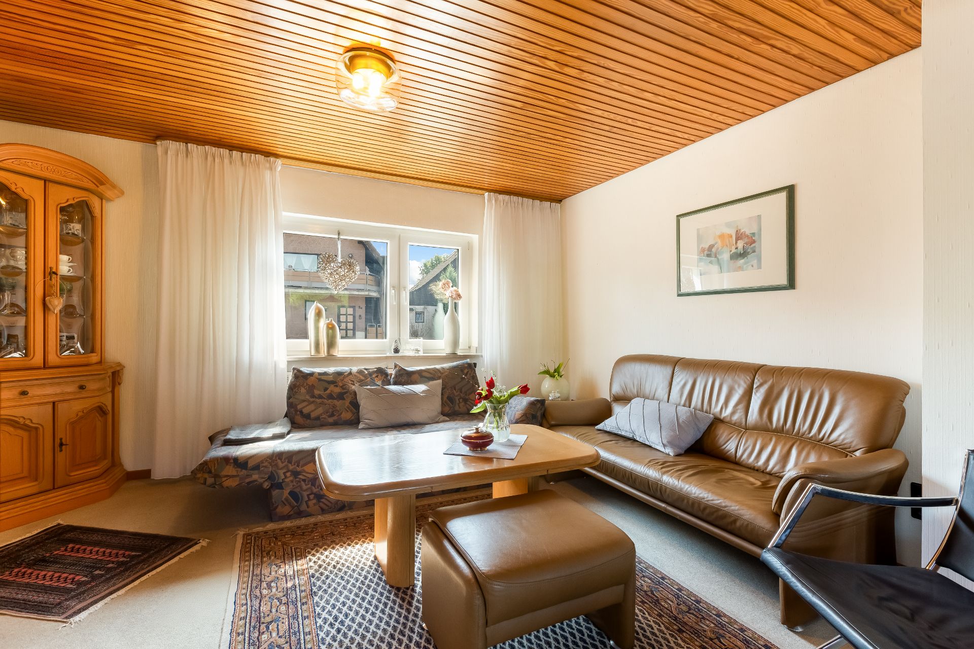 Wohnzimmer mit braunem Ledersofa, Sessel, Holztisch, Teppich und Vitrine vor Fenster mit Vorhängen.