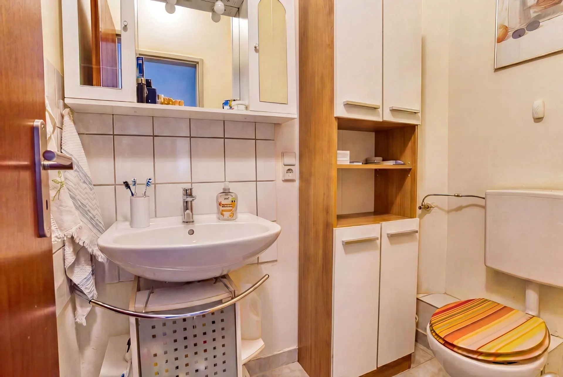 Kleines Badezimmer mit Waschbecken, Spiegelschrank, Schränken und WC mit gestreiftem Toilettensitz.
