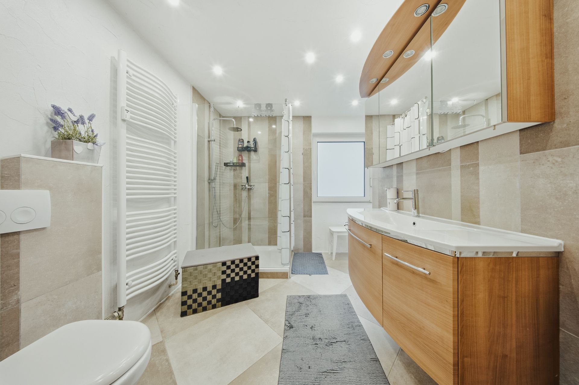 Modernes Badezimmer mit Dusche, großem Spiegel, Holzschränken und Handtuchheizung.