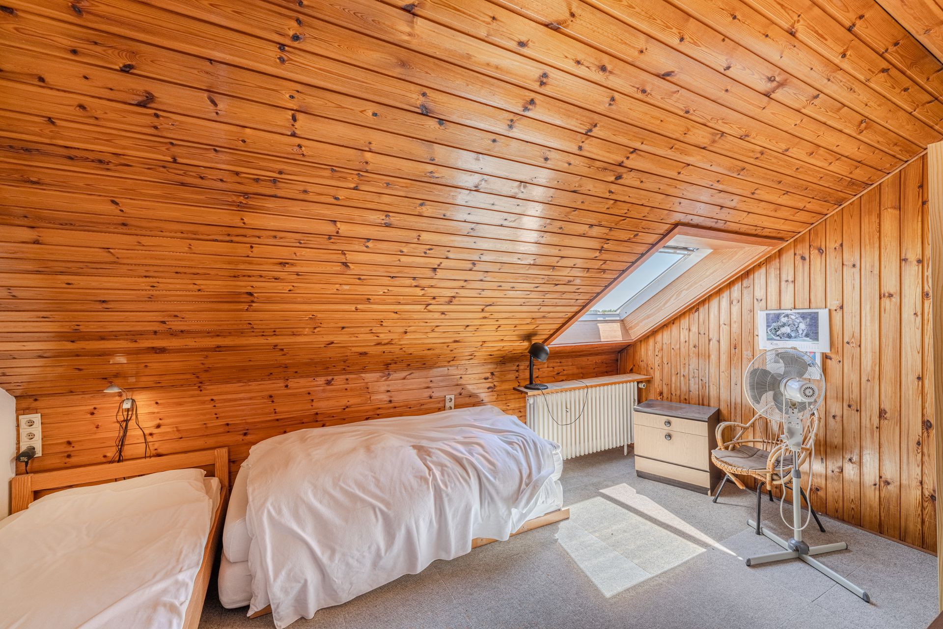 Dachzimmer mit Holzverkleidung, zwei Einzelbetten, Fenster, Stuhl, Ventilator und Nachttisch.