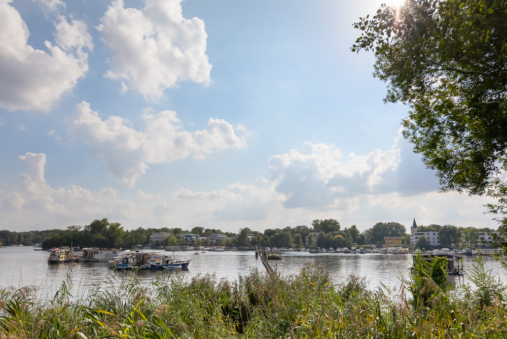Blick auf einen Fluss mit Booten, Uferbewuchs und Häusern im Hintergrund bei Sonnenschein.