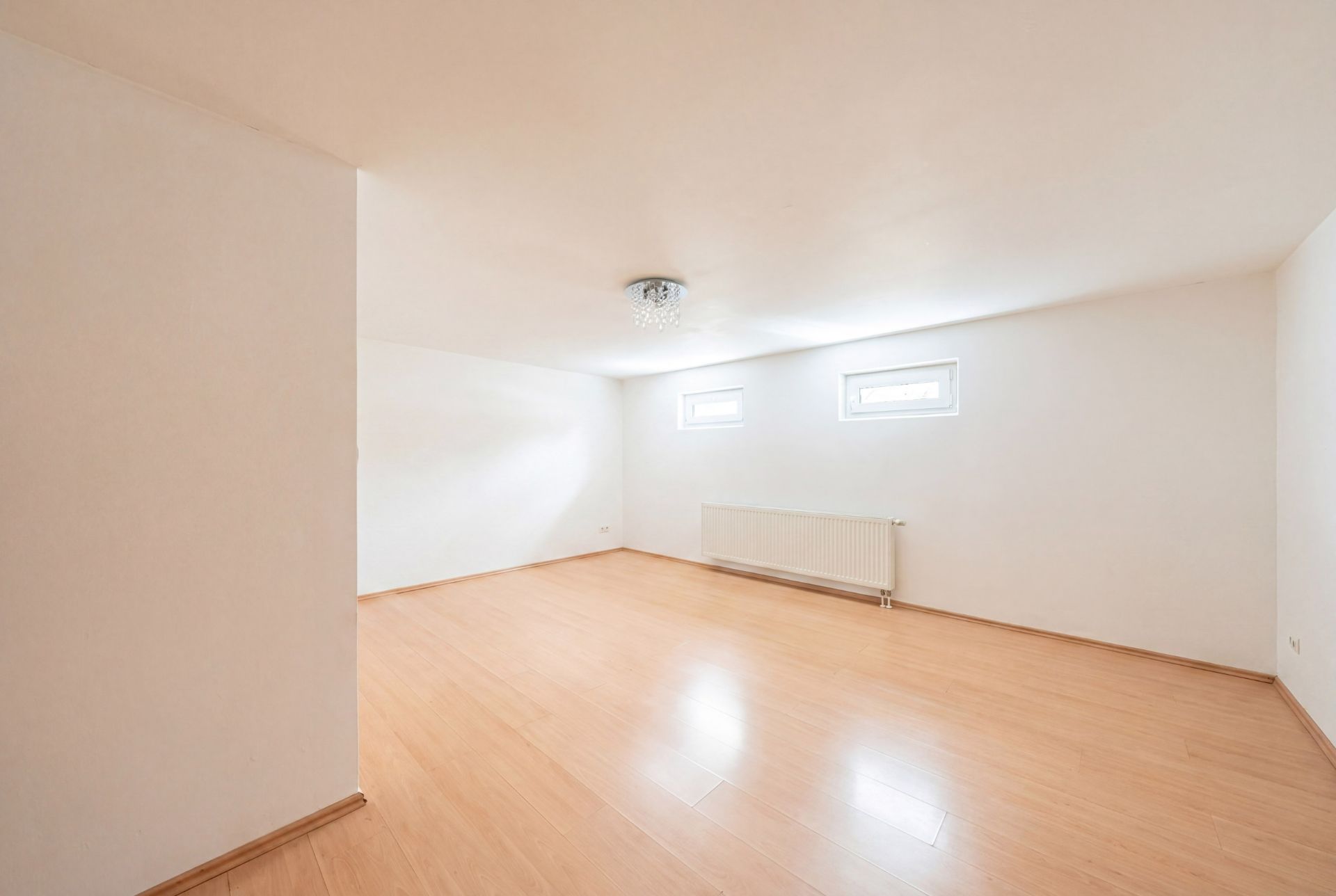 Leerer, heller Raum mit Laminatboden, zwei kleinen Fenstern und einem Heizkörper an der Wand.
