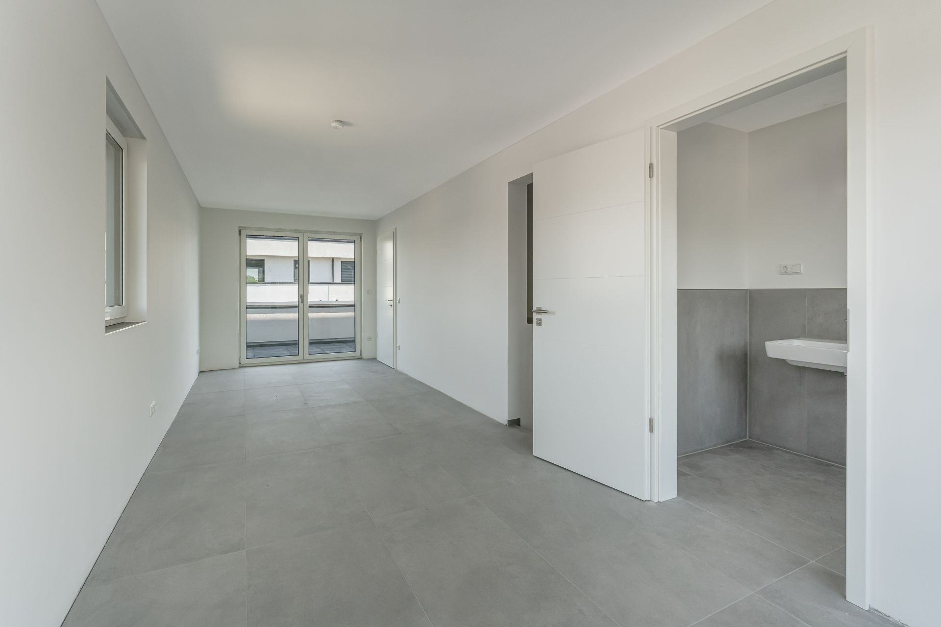 Heller, leerer Raum mit Fliesenboden, Fenster, Türen und angrenzendem Badezimmer.