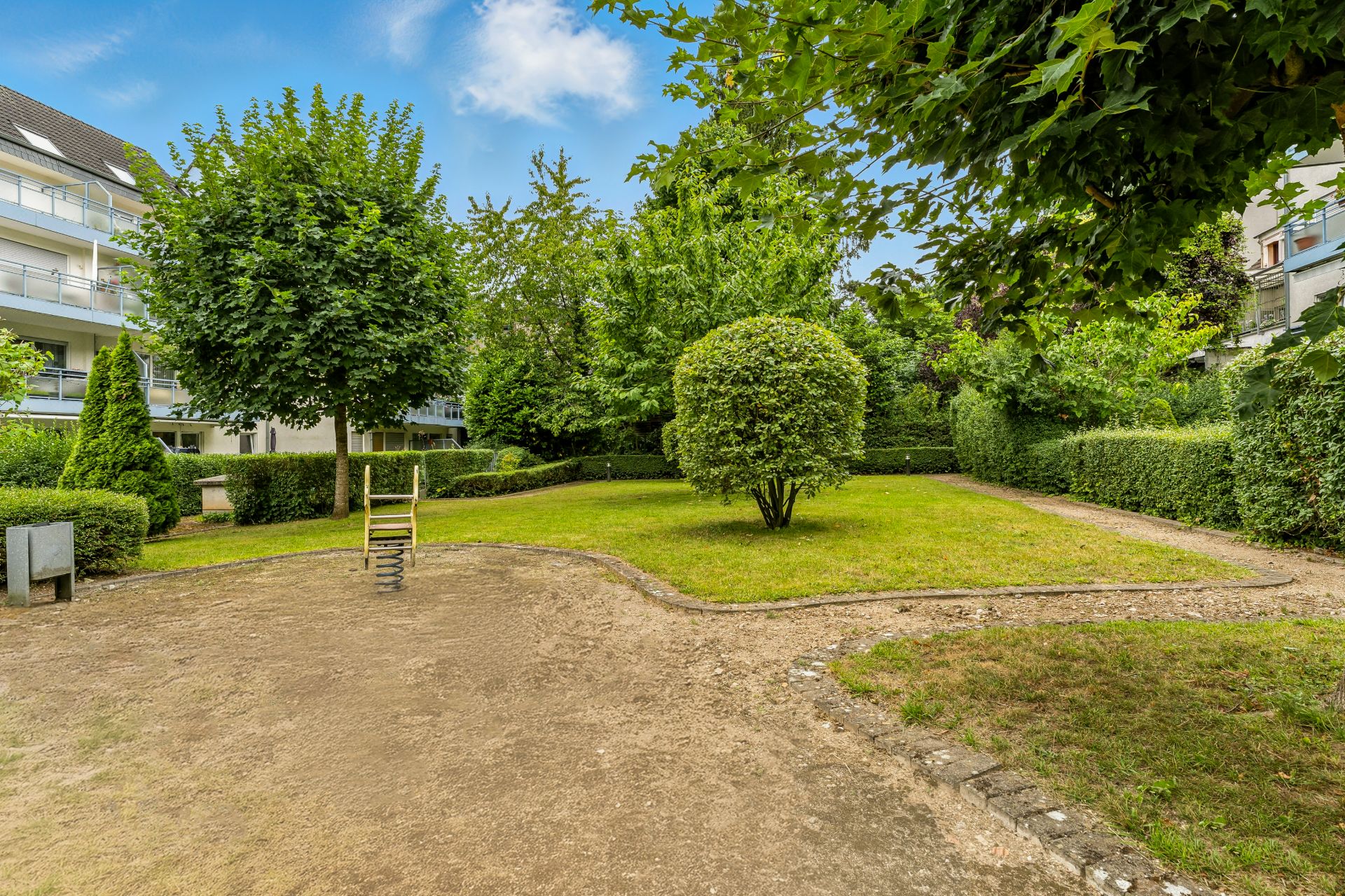 Gepflegter Garten mit Rasen, Bäumen, Hecken und einem kleinen Spielgerät im Vordergrund.