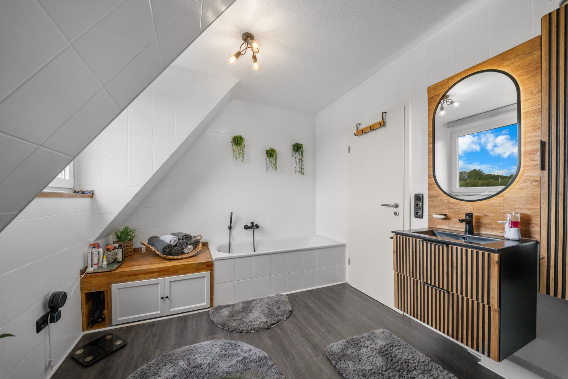 Modernes Badezimmer mit Badewanne, Holzmöbeln, Spiegel und drei Teppichen unter Dachschräge.