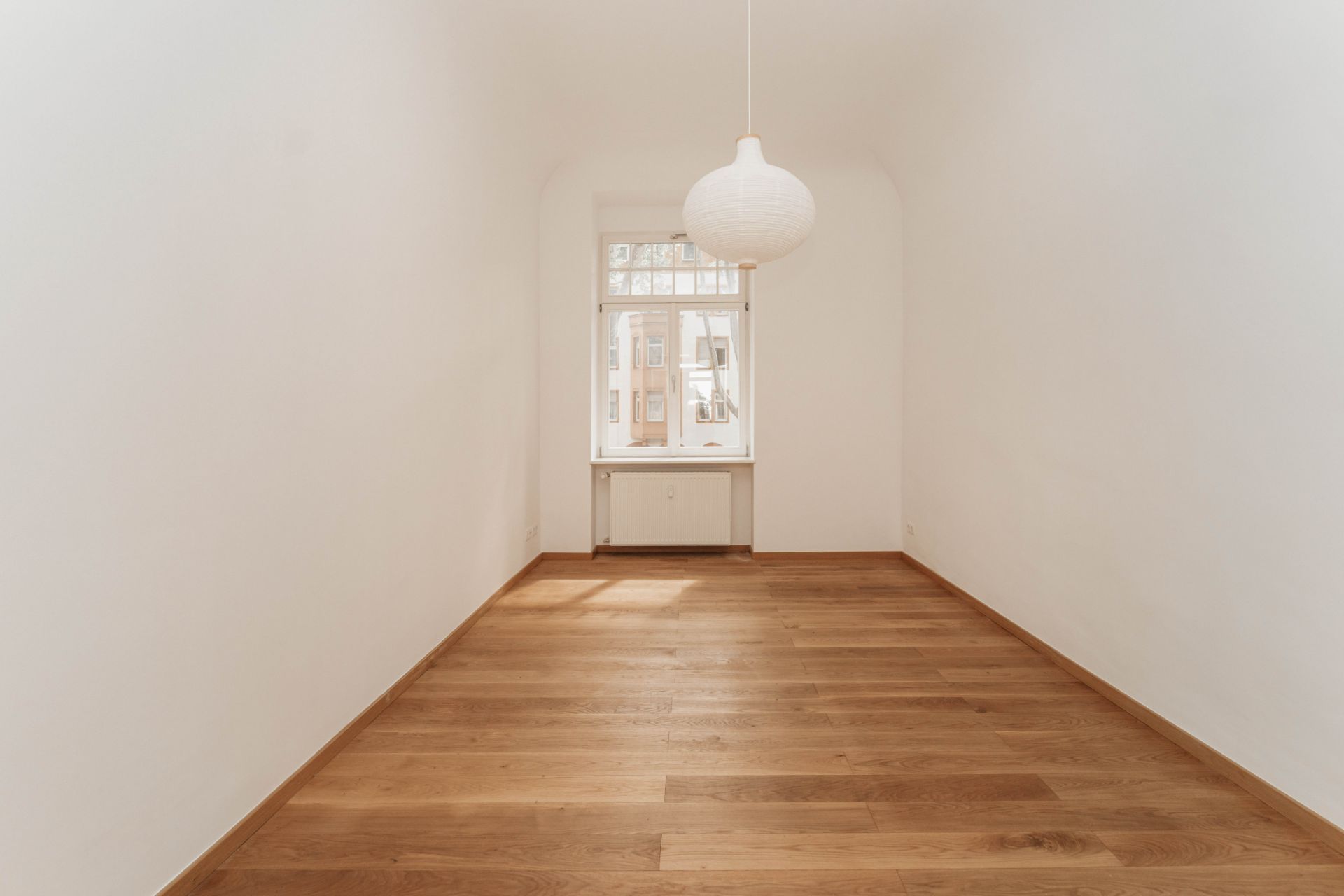 Leerer Raum mit Holzfußboden, weißer Wand und Fenster, Deckenlampe hängt in der Mitte.