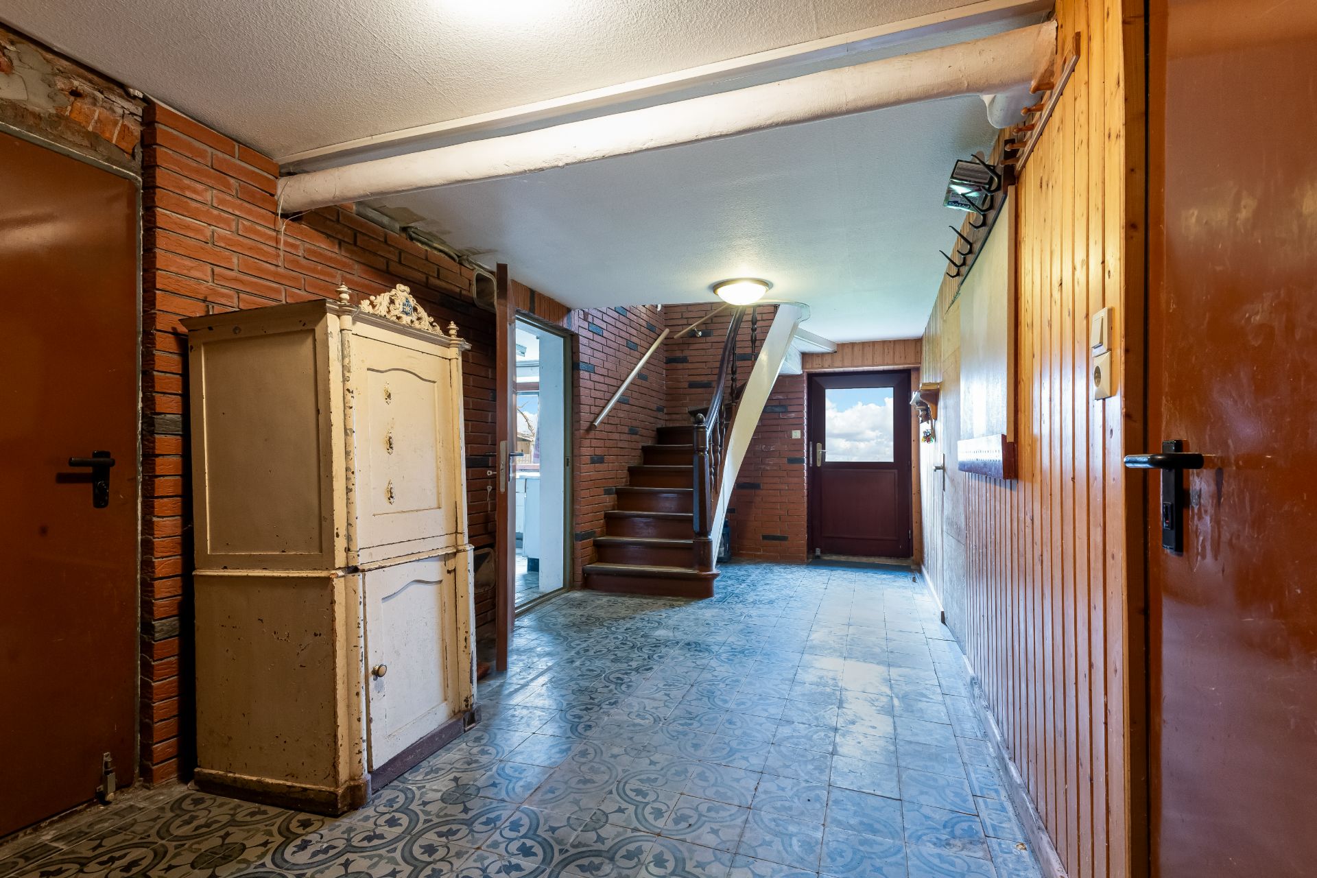 Flur mit gemustertem Boden, altem Schrank, Treppe und Tür mit Fenster nach draußen.