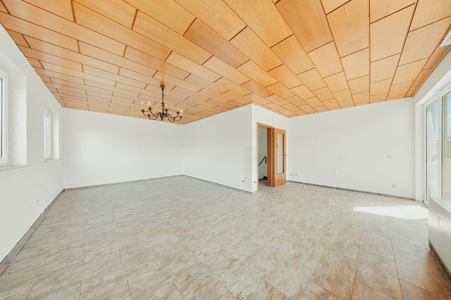 Heller, leerer Raum mit Fliesenboden, Holzdecke, Kronleuchter und mehreren Fenstern.