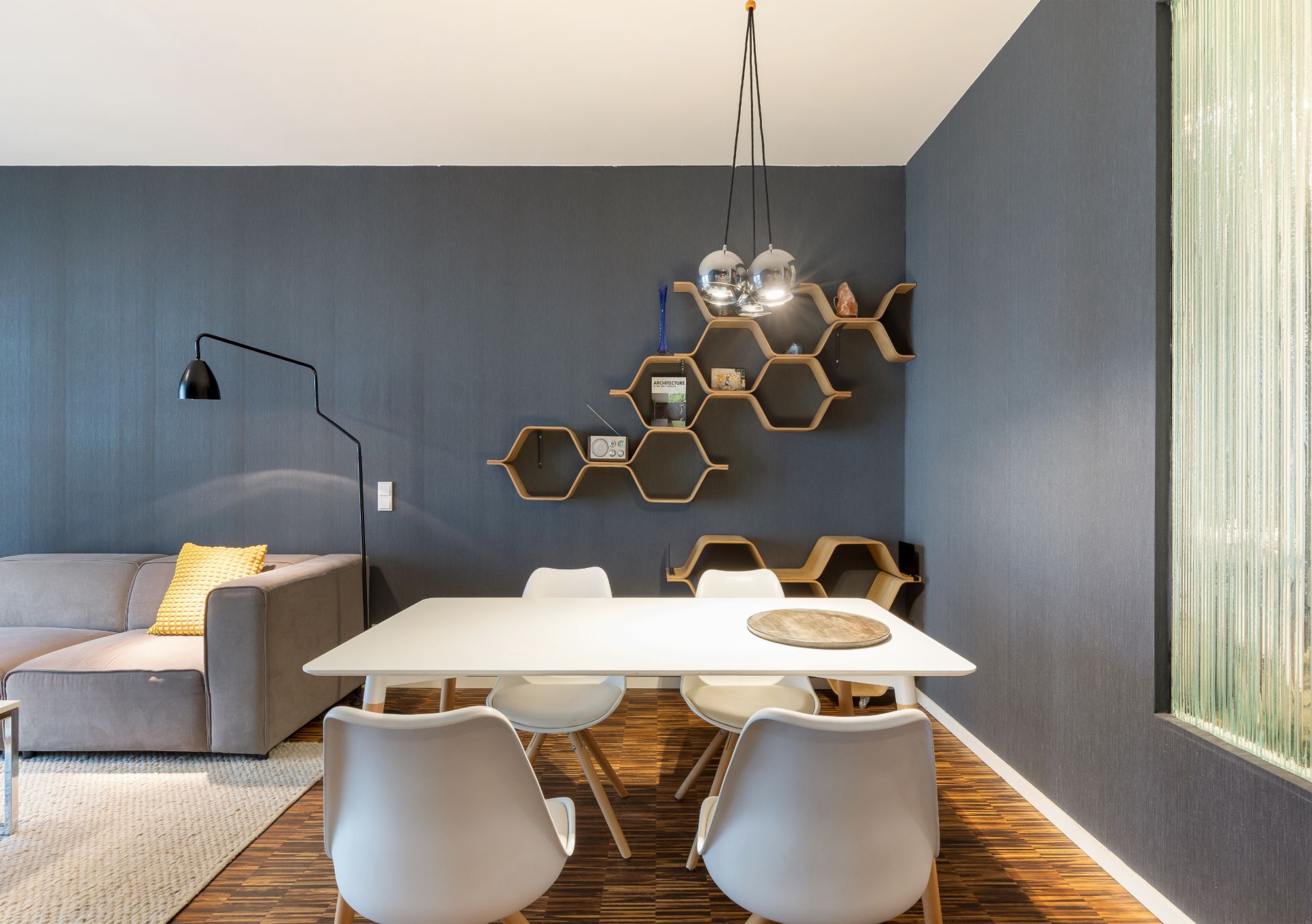 Essbereich mit weißem Tisch, vier Stühlen, grauem Sofa und Wabenregalen an blauer Wand.