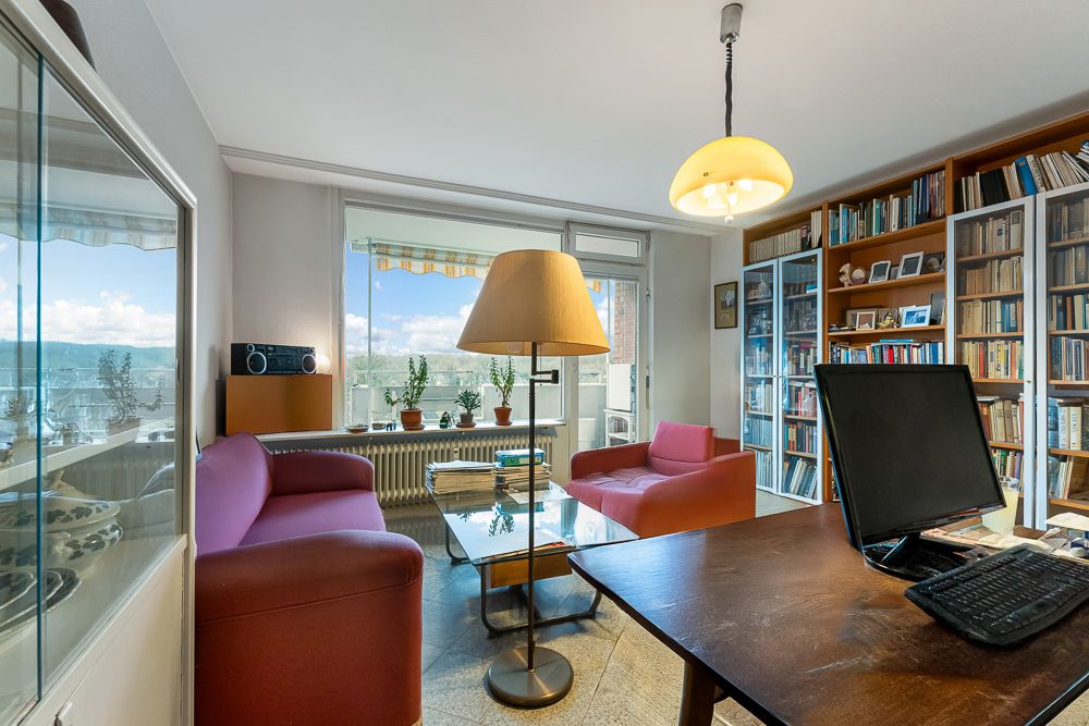 Wohnzimmer mit zwei roten Sesseln, Bücherregalen, Schreibtisch und Glas-Couchtisch, große Fenster im Hintergrund.
