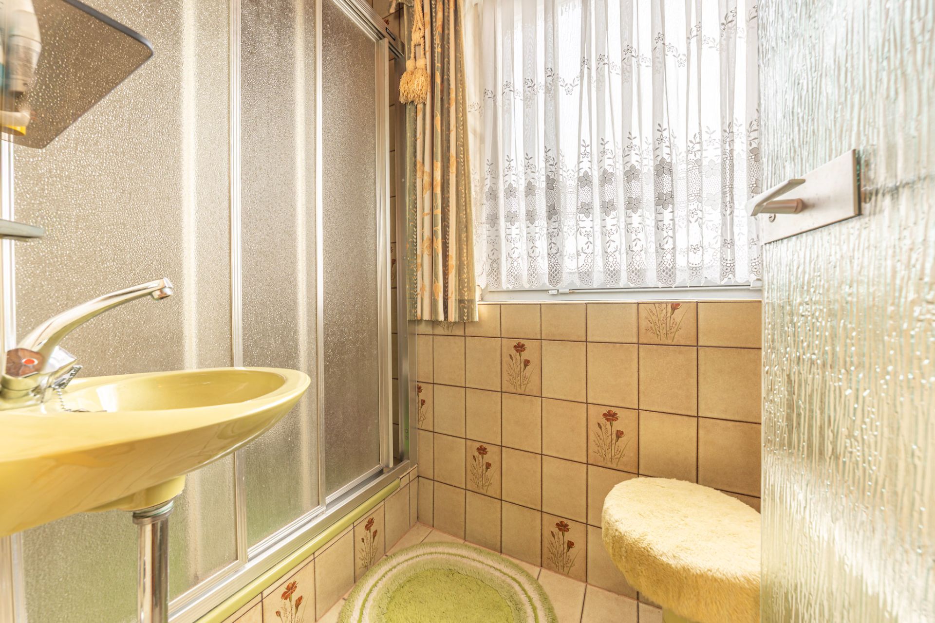Kleines Badezimmer mit gelbem Waschbecken, Dusche, WC und Fenster mit Gardine.
