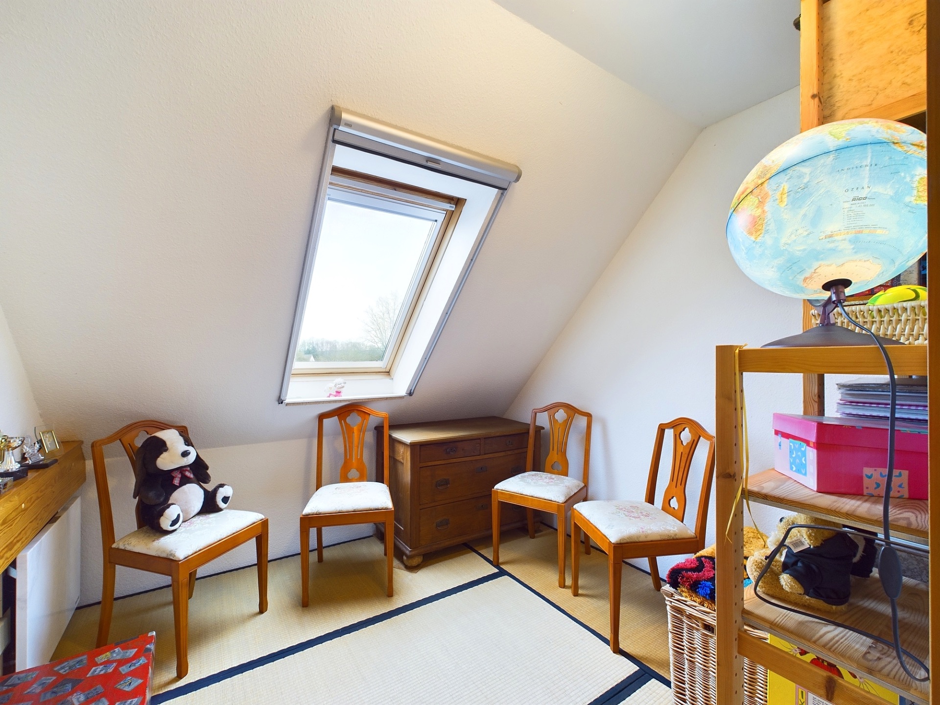 Kleines Zimmer mit Dachfenster, drei Stühlen, einer Kommode und einem Regal mit Globus.