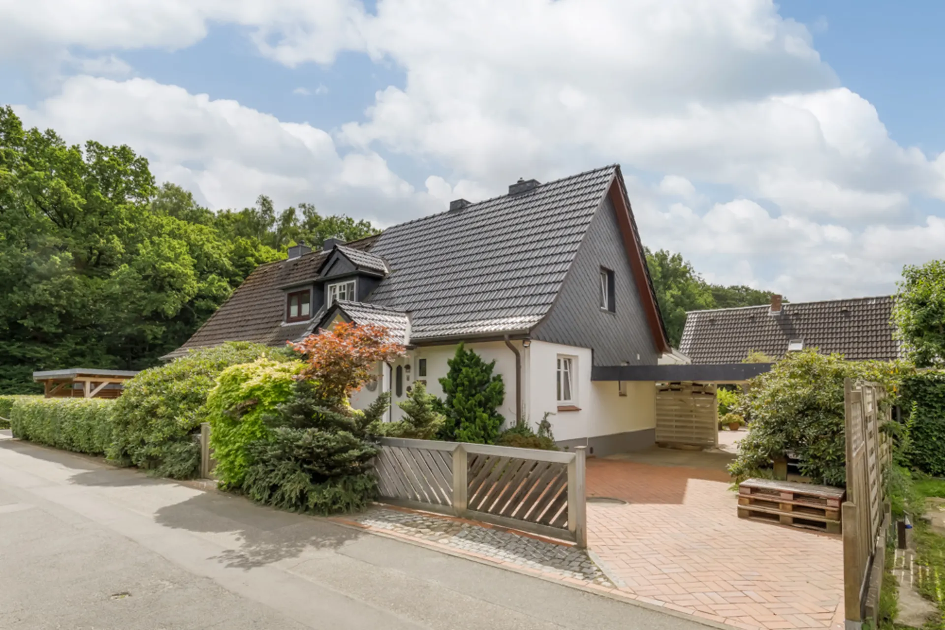 Einfamilienhaus mit dunklem Dach, Vorgarten und Einfahrt mit Tor, umgeben von viel Grün.