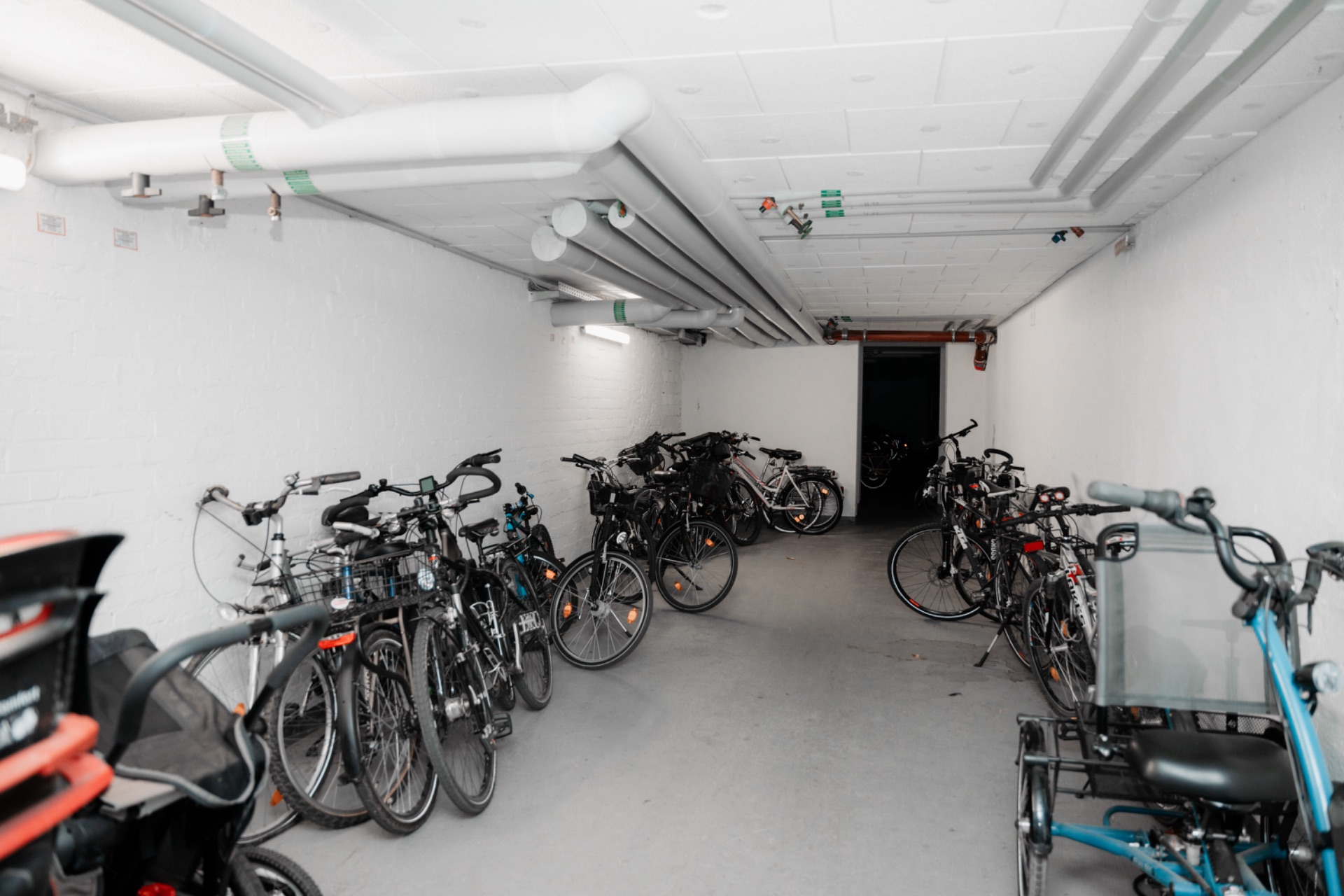 Fahrradraum mit mehreren abgestellten Fahrrädern in einem Keller mit weißen Wänden und Rohren an der Decke.