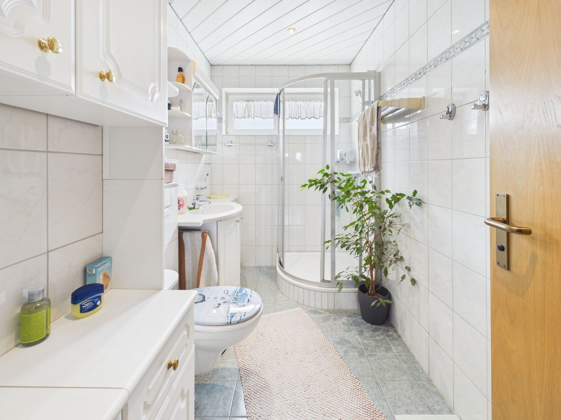 Helles Badezimmer mit Dusche, Waschbecken, WC, Schränken und einer Pflanze im Topf.