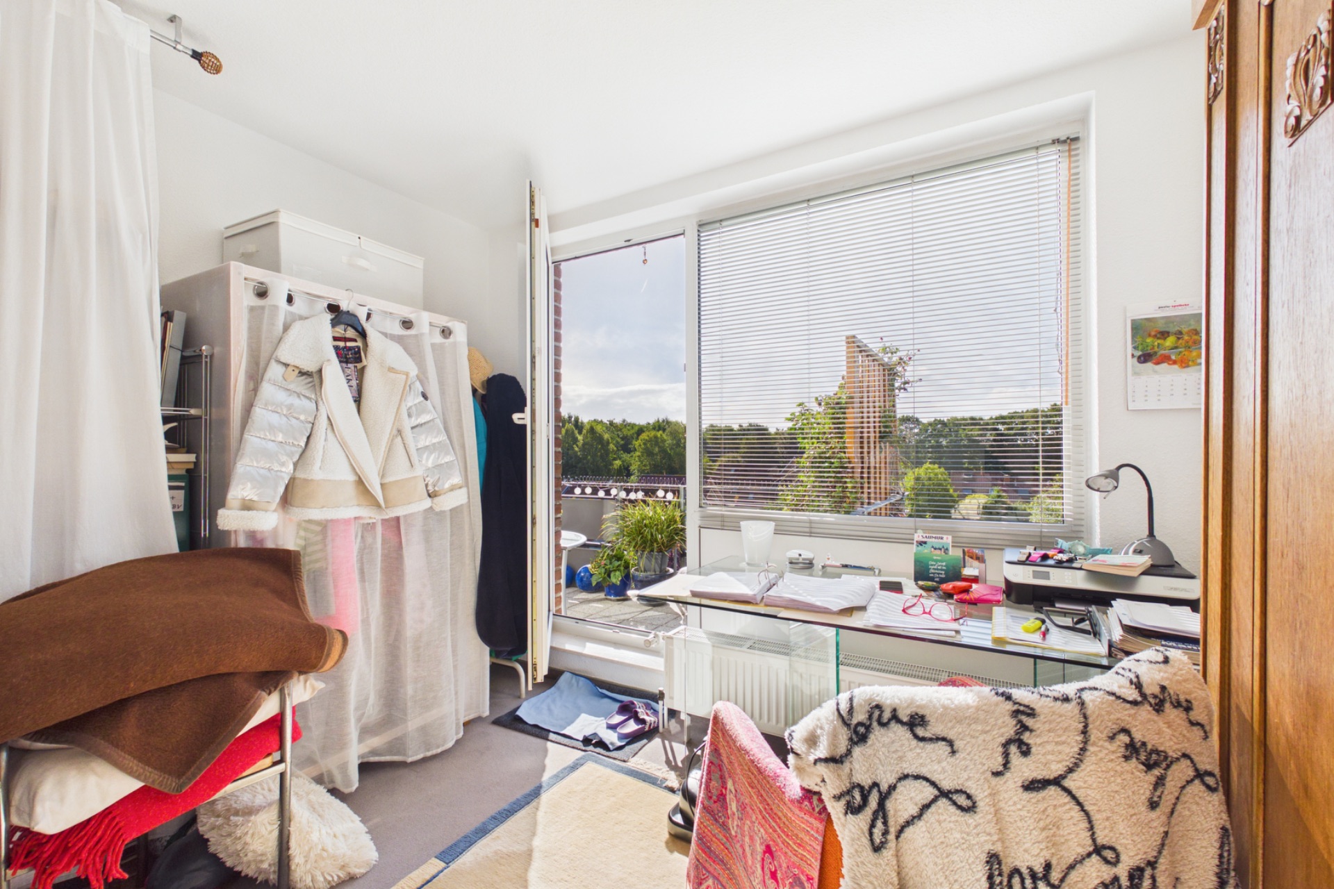 Helles Zimmer mit großem Fenster, Schreibtisch, Stuhl und Kleidung auf einem Garderobenständer.