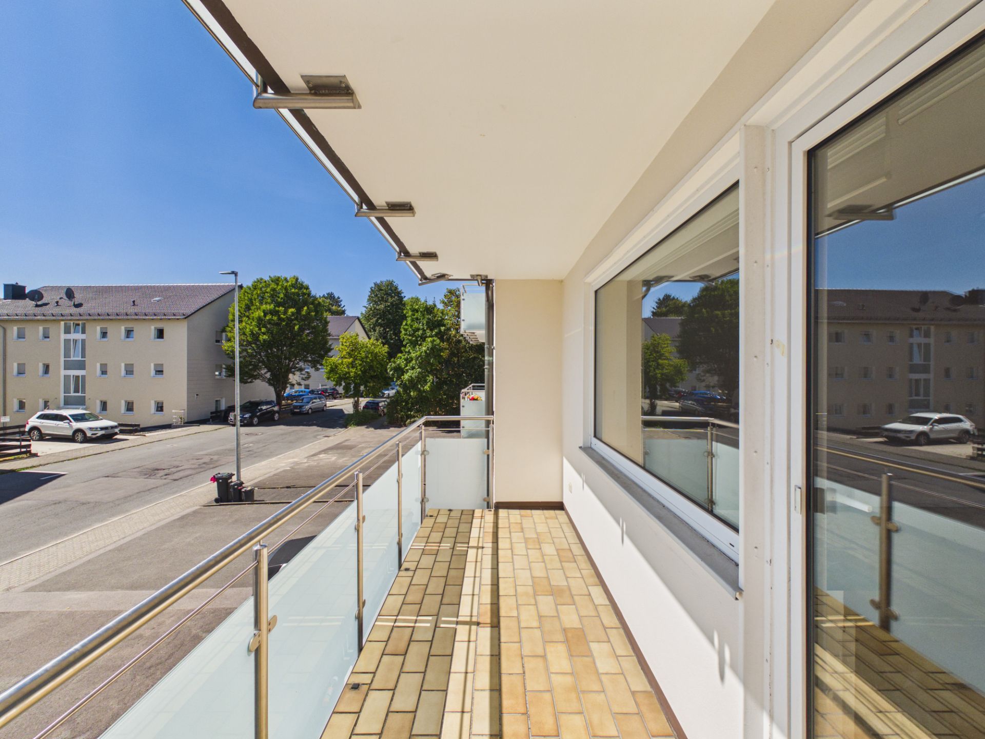 Balkon mit Glasgeländer und Fliesenboden, Blick auf Straße und Wohnhäuser.