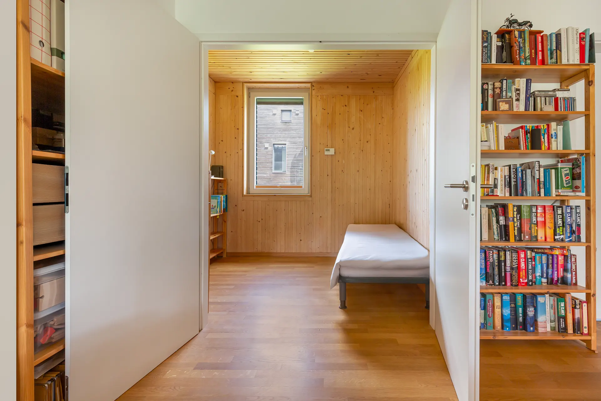 Kleines Zimmer mit Holzverkleidung, Einzelbett, Fenster und Bücherregal.