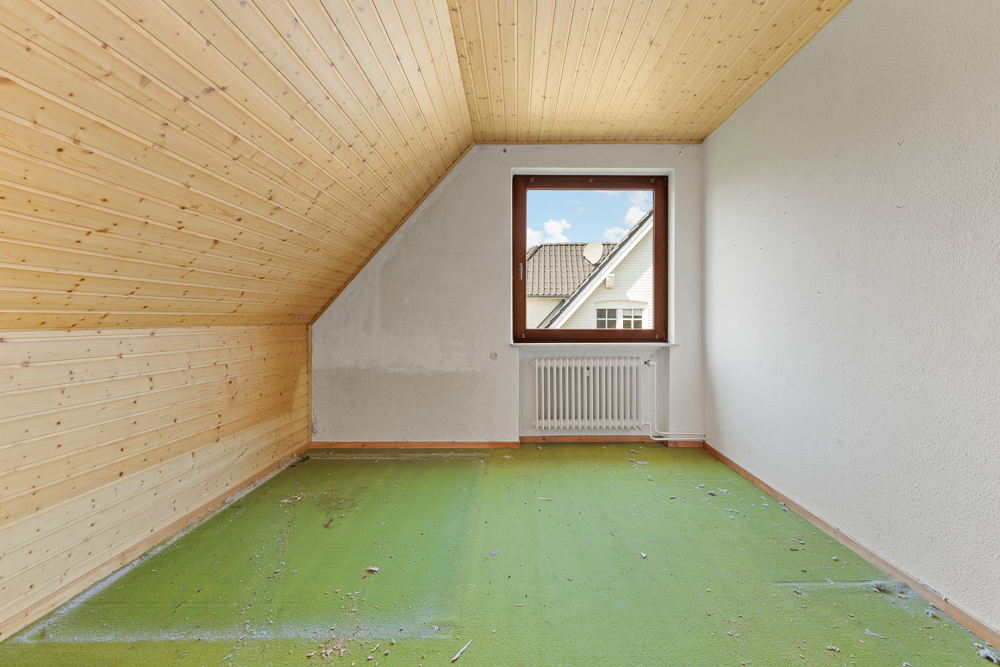 Leeres Zimmer mit Holzdecke, grünem Teppichboden und Fenster mit Blick auf Dach.