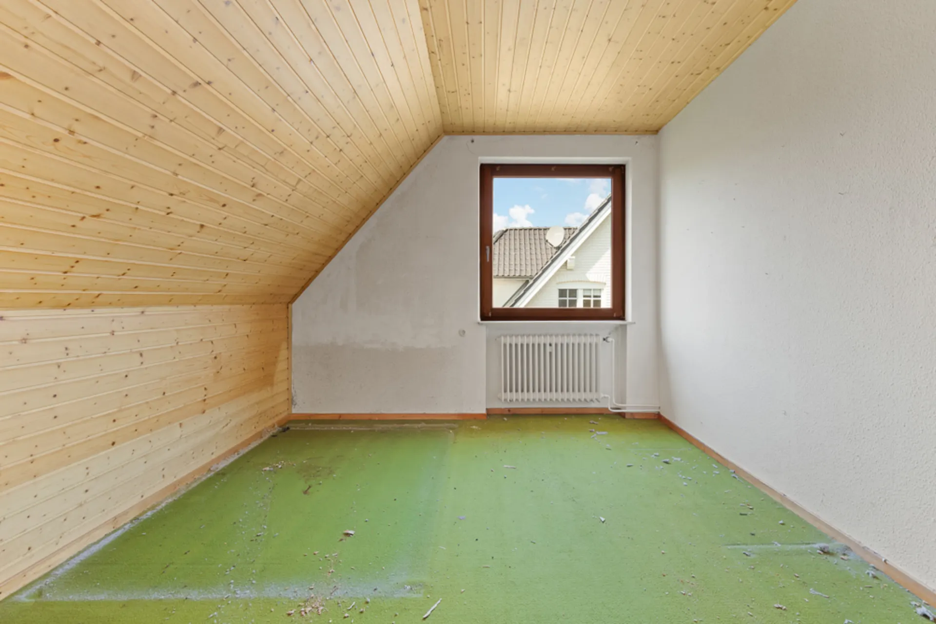 Leeres Zimmer mit Holzdecke, grünem Teppichboden und Fenster mit Blick auf Dach.