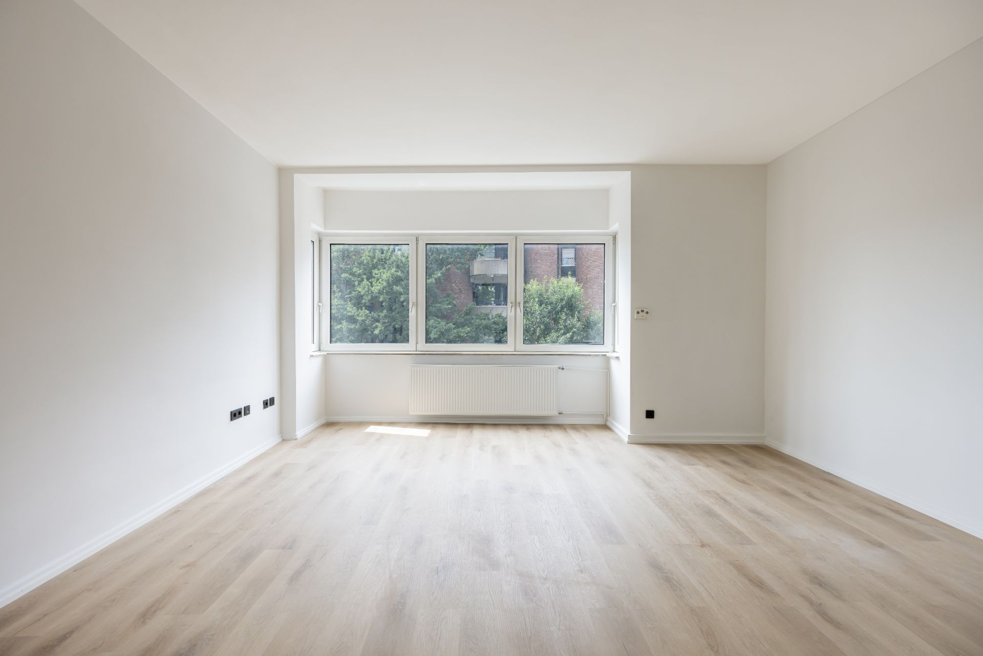 Leerer, heller Raum mit Holzboden und großem Fenster mit Blick ins Grüne.