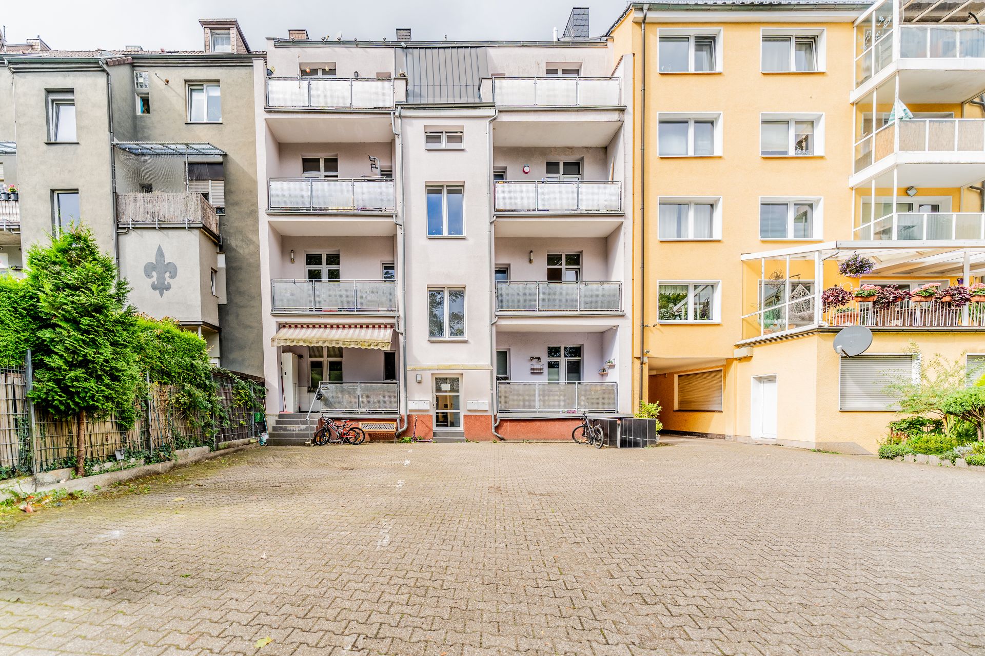 Mehrfamilienhaus mit Balkonen und einem gepflasterten Hof, mehrere Fahrräder vor dem Eingang.