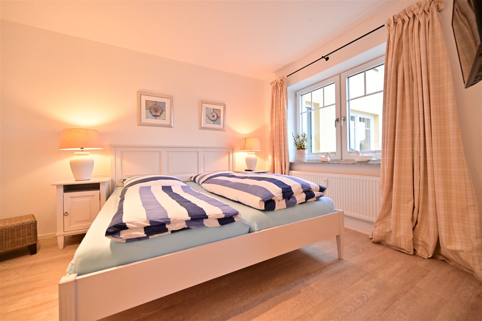 Helles Schlafzimmer mit Doppelbett, gestreifter Bettwäsche, Nachttischen und Fenster mit Vorhängen.