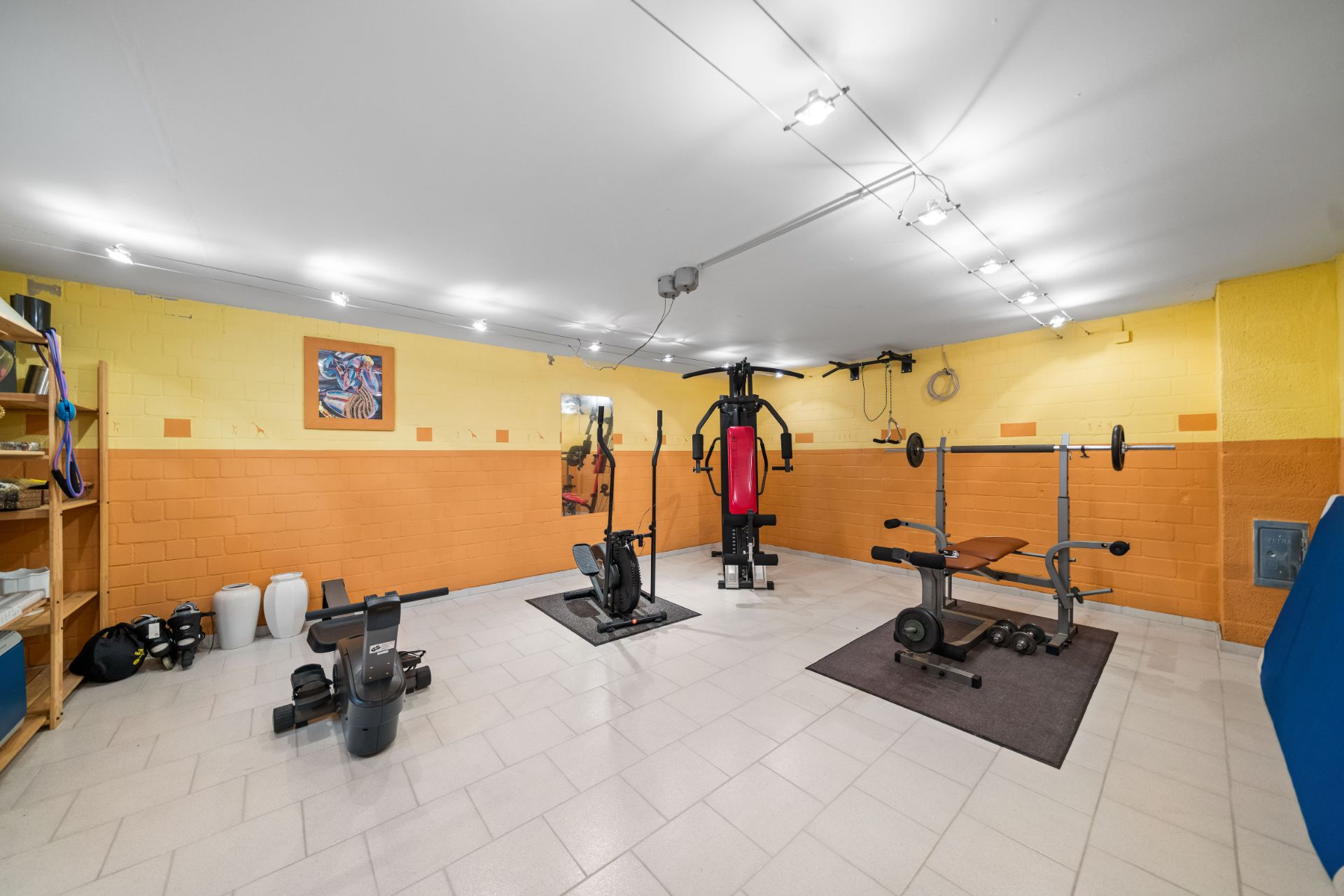 Fitnessraum mit Trainingsgeräten, Hanteln, Regal und hellem Fliesenboden.
